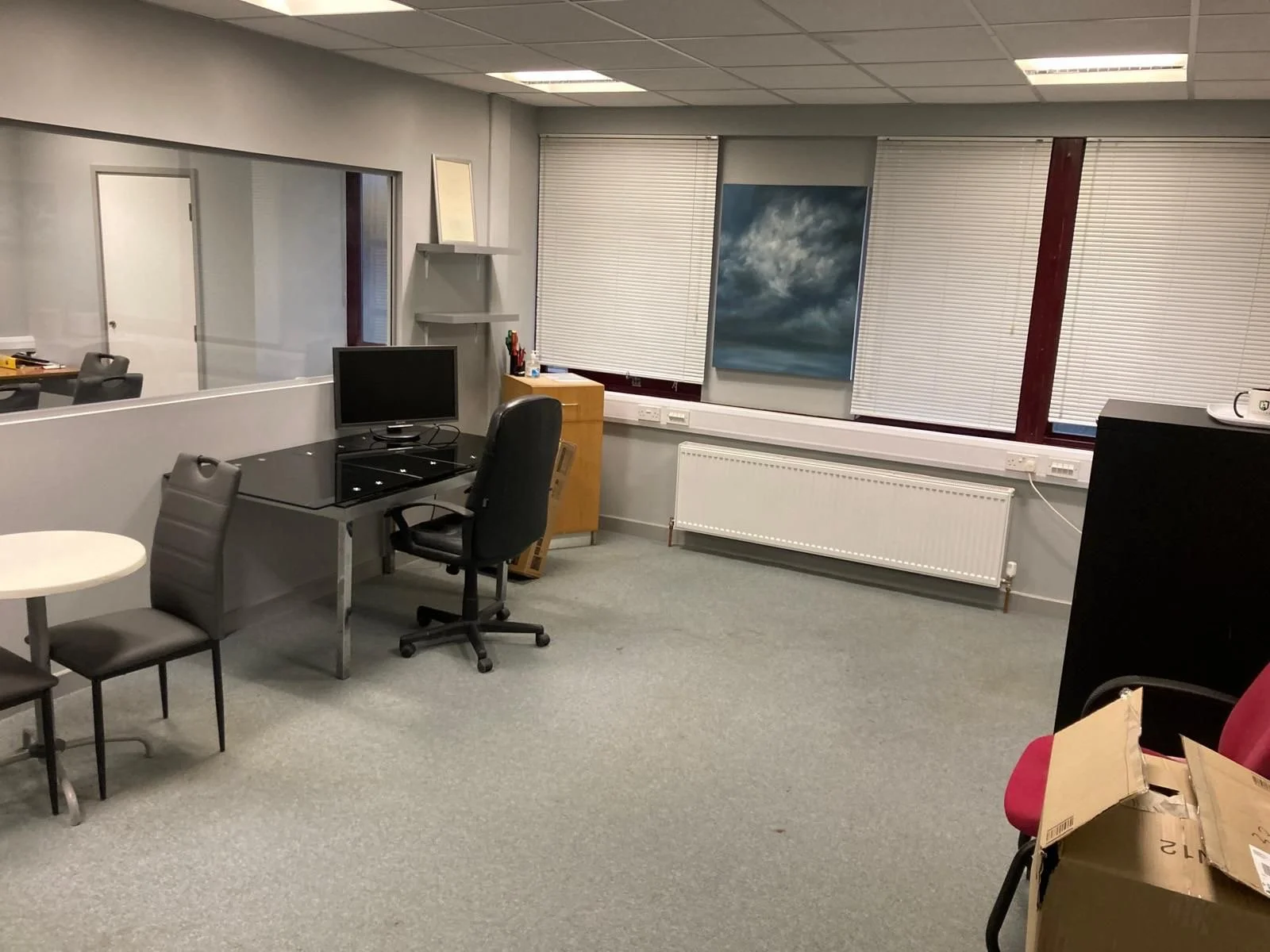 An office with gray carpet, several chairs, a small desk with a computer monitor, blinds on the windows, a radiator, a painting of a cloudy sky, and various office supplies.