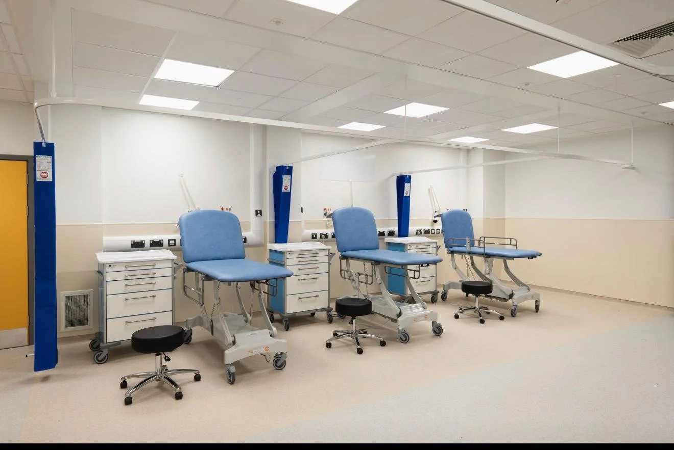 Empty medical treatment room with four blue reclining patient chairs, each with white cabinets and black stools on wheels.