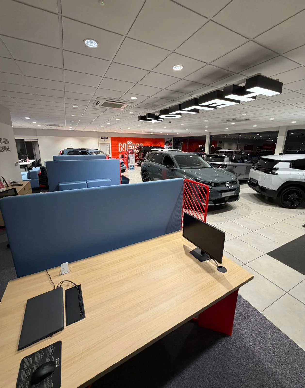 View of a car dealership showroom with several new cars on display, a seating area with blue chairs, a desk with a computer, and a neon sign that says 'NEW'.