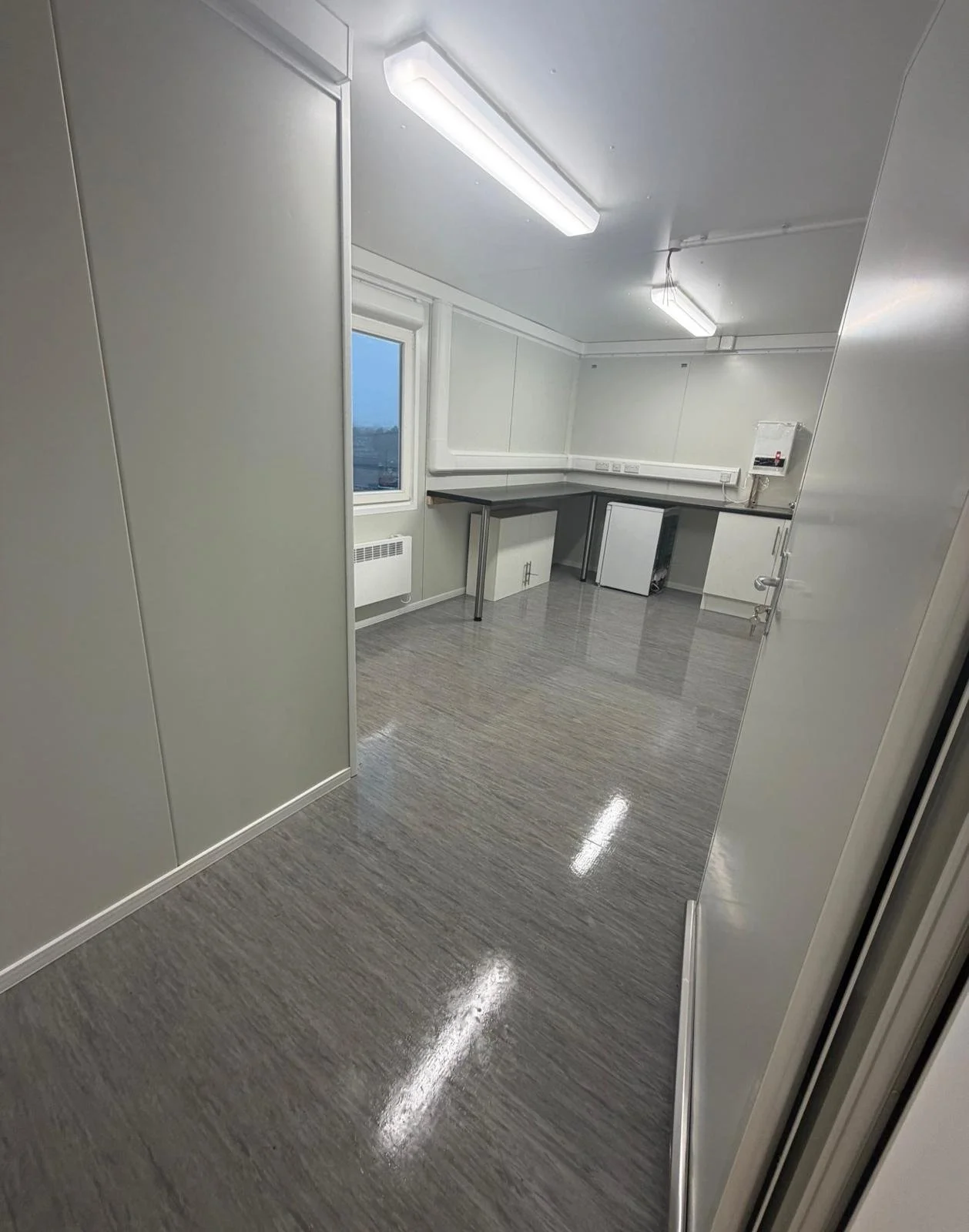 Empty room with gray flooring, white walls, fluorescent lighting, a window, a radiator, and a small counter.