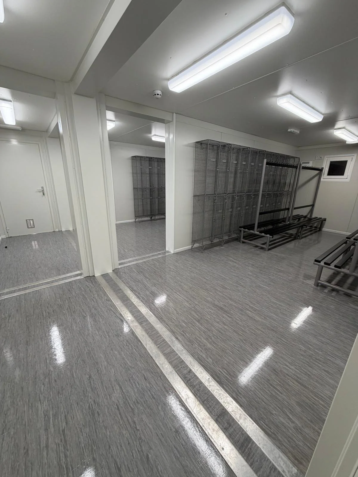 Empty locker room area with metal benches, storage lockers, bright fluorescent lighting, and a small window.