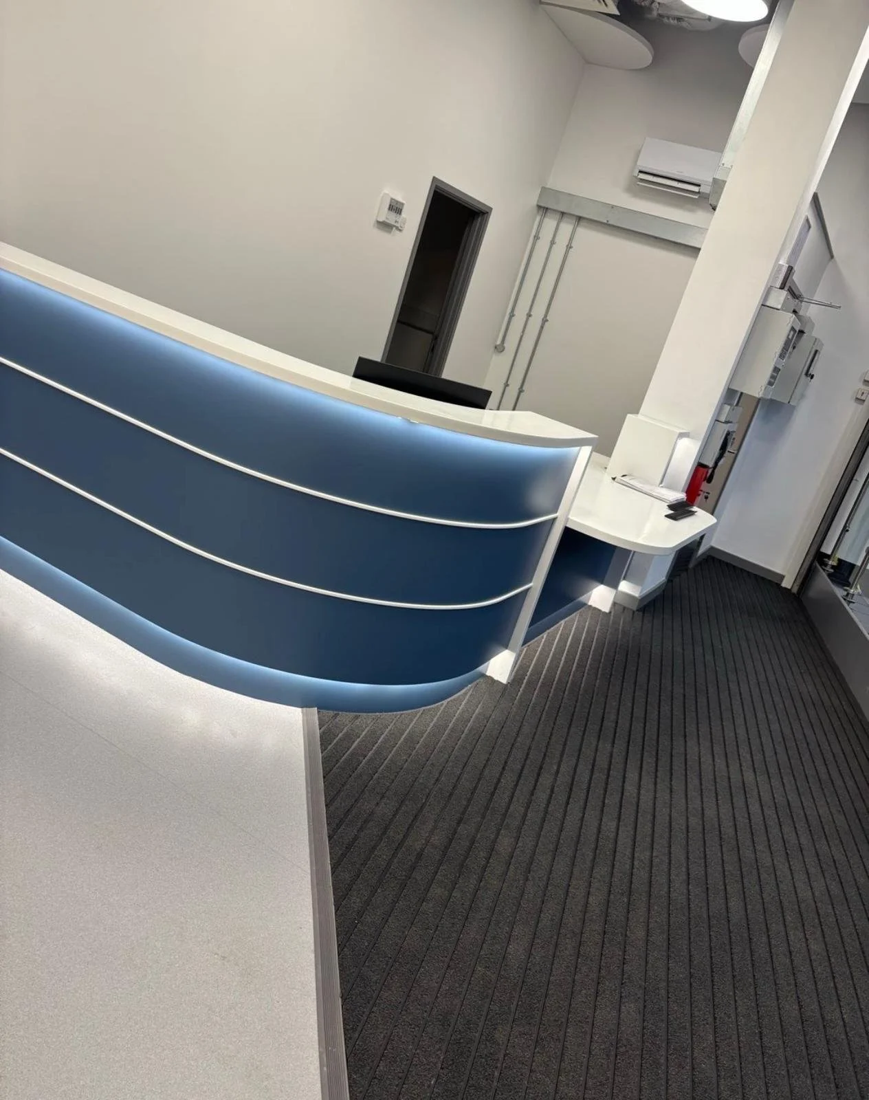 Modern reception desk with blue curved panels and backlit lighting, situated in a sleek, minimalistic office or medical facility with gray carpeting and white walls.
