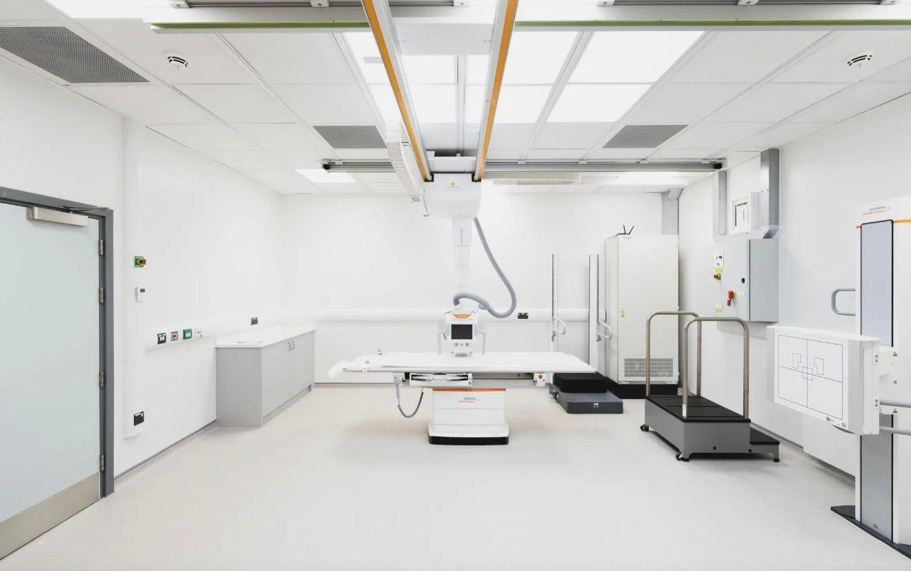 A bright, sterile medical imaging room with an X-ray machine and patient table, surrounded by white walls and medical equipment.