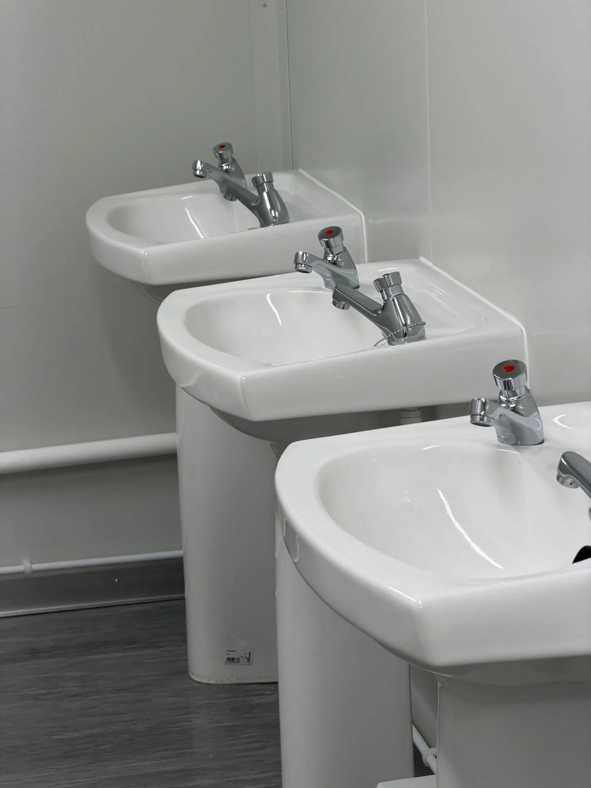 Three white sinks with chrome faucets in a restroom, positioned in front of a plain wall with a mirror above them.