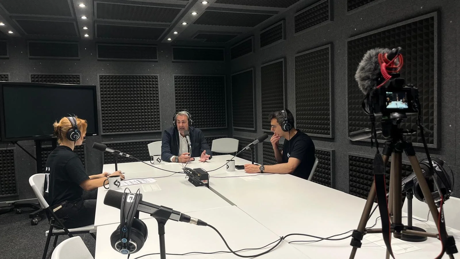 Tres personas en un estudio de grabación en una mesa, con grabadoras de voz y micrófonos, discutiendo para un programa de radio o podcast.