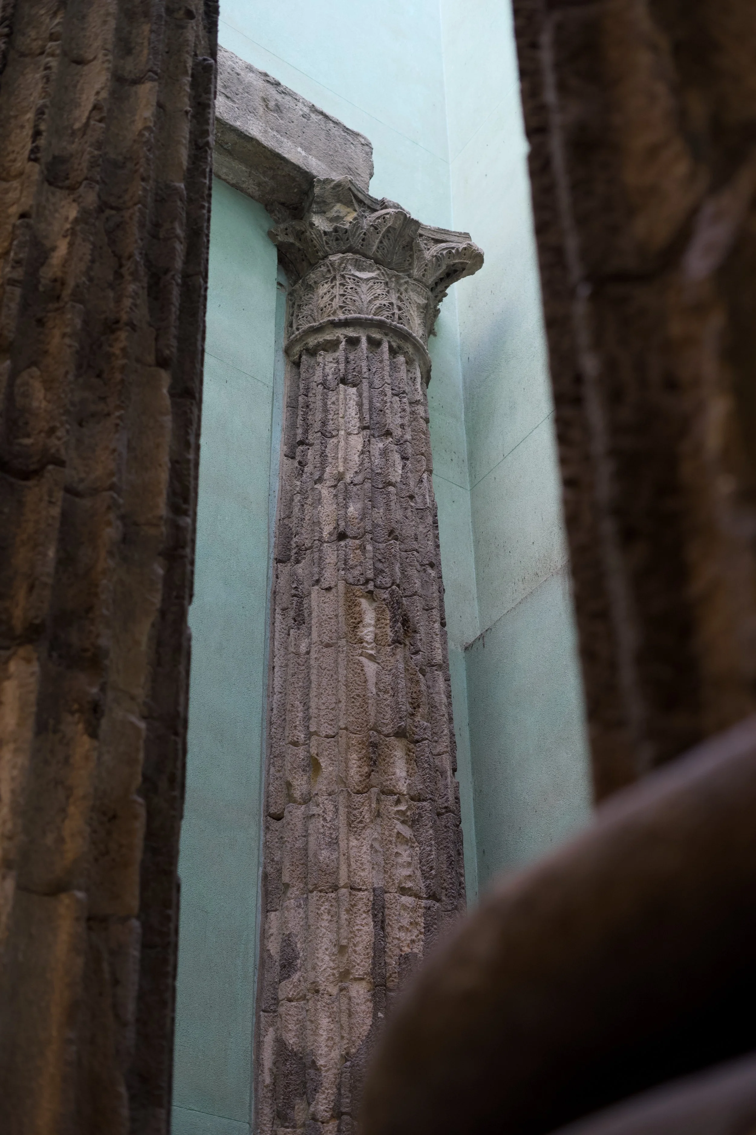 Columna antigua de piedra con detalles decorativos en la parte superior, vista entre muros de ladrillo y pared de color mint en un edificio antiguo.