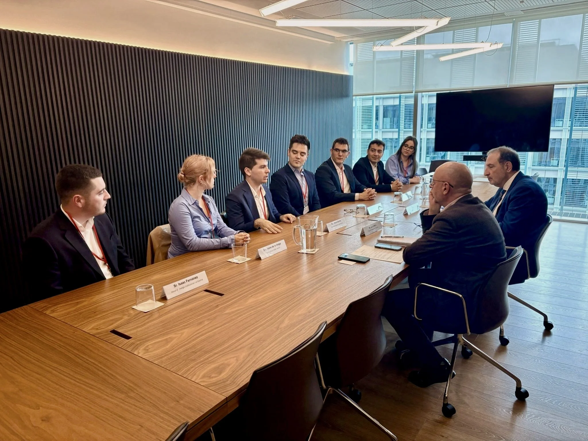 Grupo de personas sentadas en una mesa de reunión en una sala moderna con ventanas grandes y vista urbana.