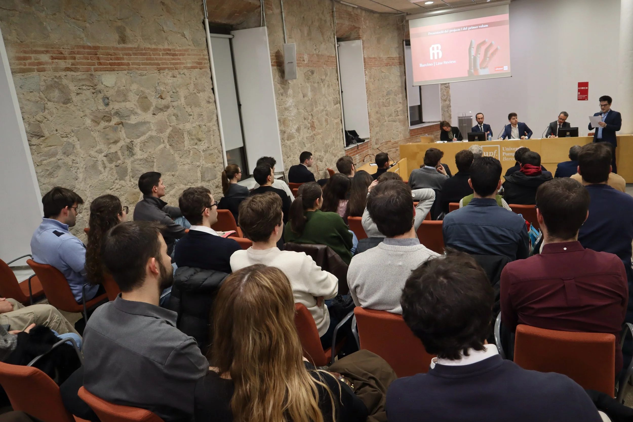 Auditorio universitario con varias personas sentadas y un ponente de pie hablando frente a una pantalla en una conferencia.