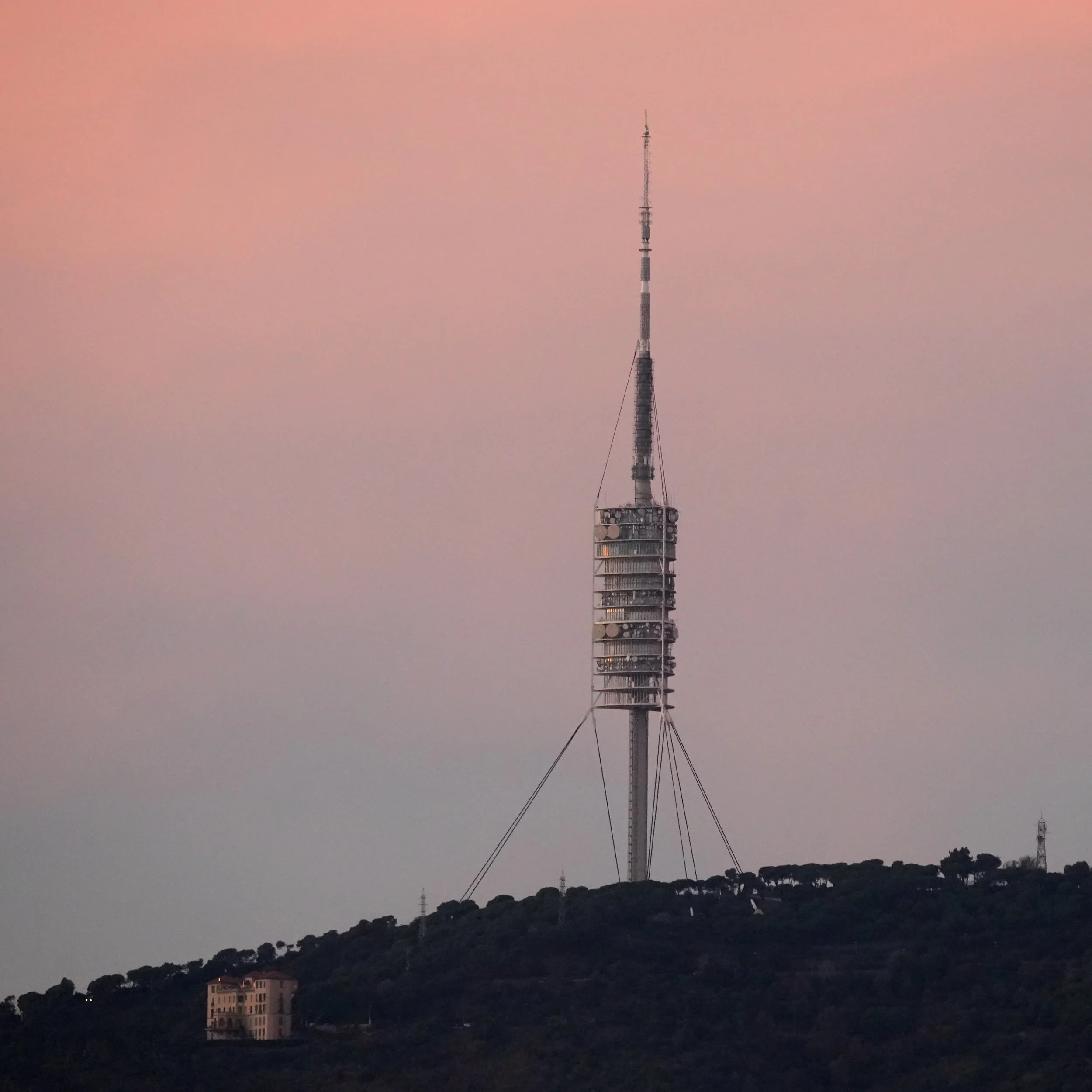 Una torre de telecomunicaciones alta en un paisaje con colinas y edificios en la base, durante el atardecer o amanecer con cielo ligeramente rosado.