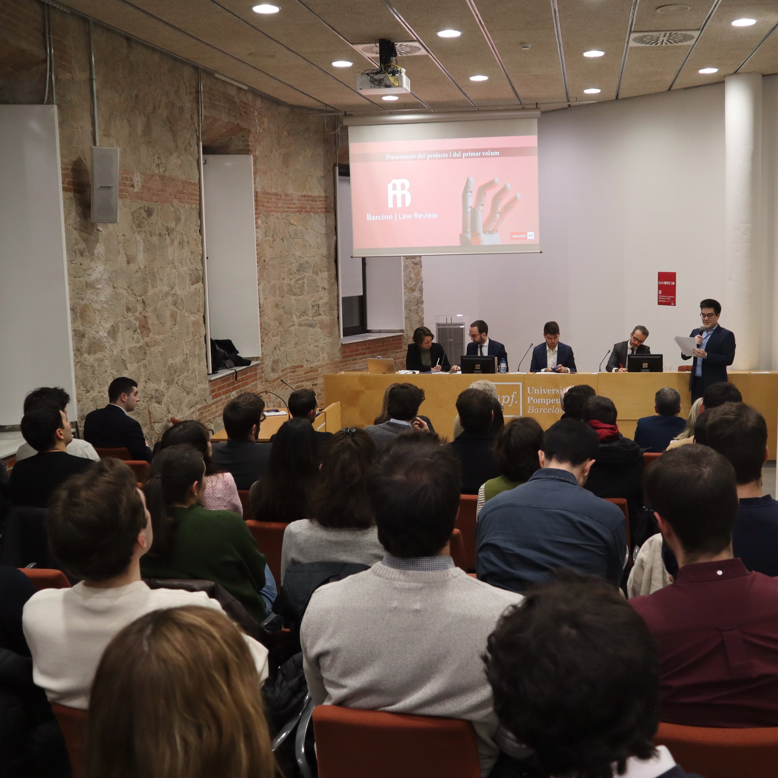 Grupo de personas asistiendo a una conferencia en un auditorio, con un proyector en el techo y una pantalla en la pared. Hay un panel de discusión con cinco hombres, uno de ellos hablando con un micrófono, mientras los demás toman notas o escuchan.
