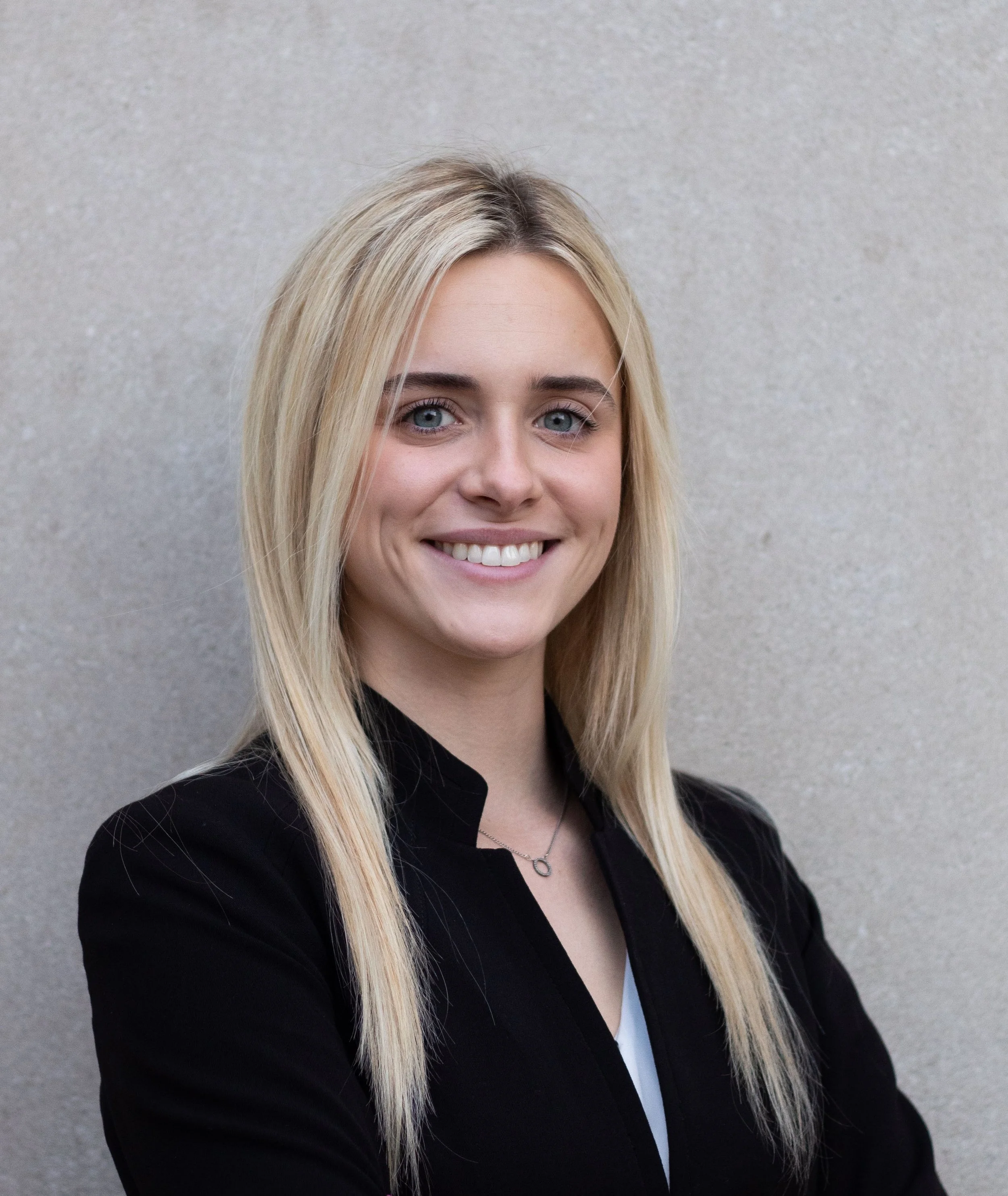 A woman with long blonde hair wearing a black blazer and a necklace, smiling in front of a plain beige wall.