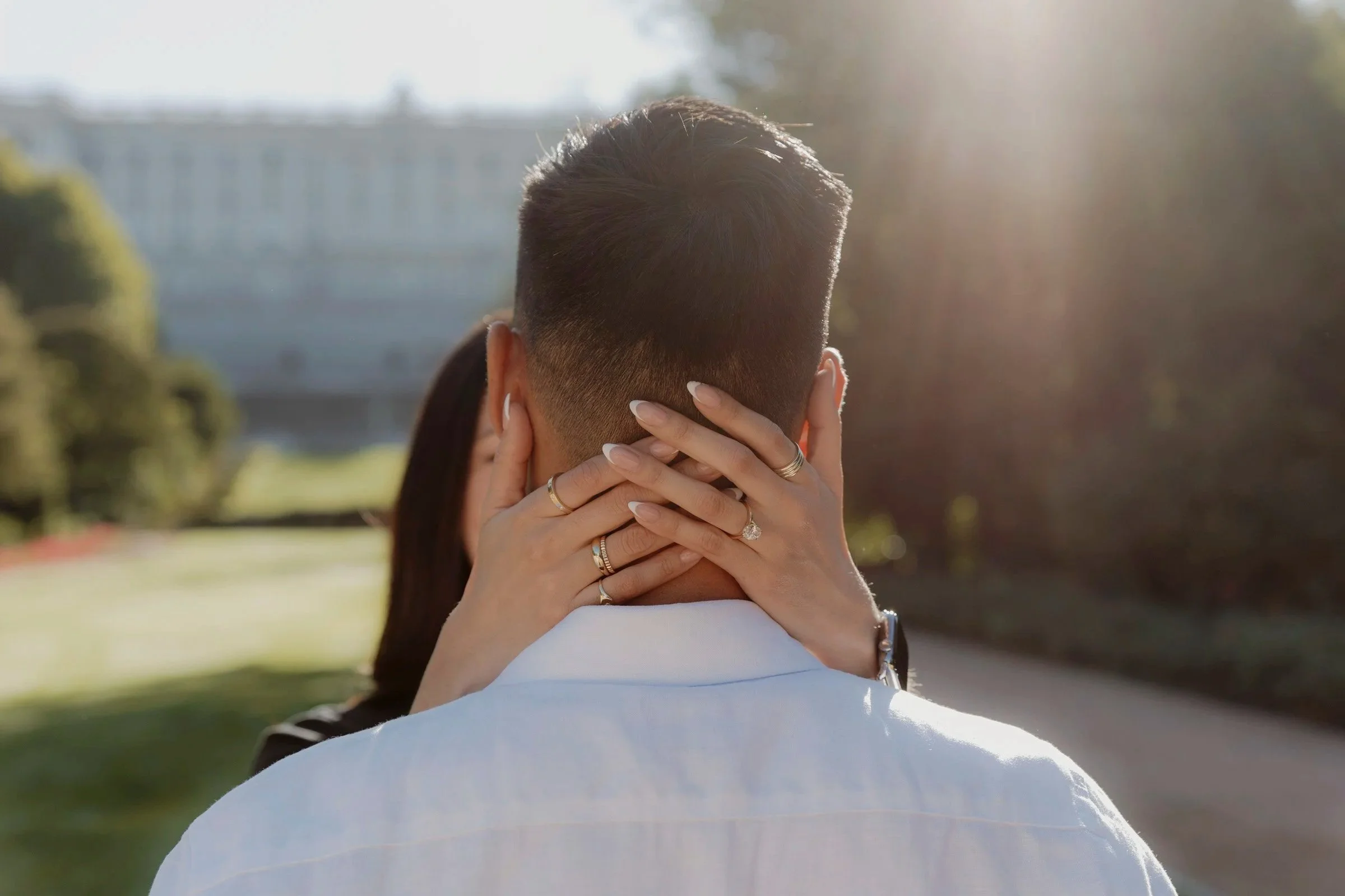 A couple is embracing outdoors, with the woman’s hands around the man’s neck, showing rings on her fingers. Sunlight creates a warm glow in the background.