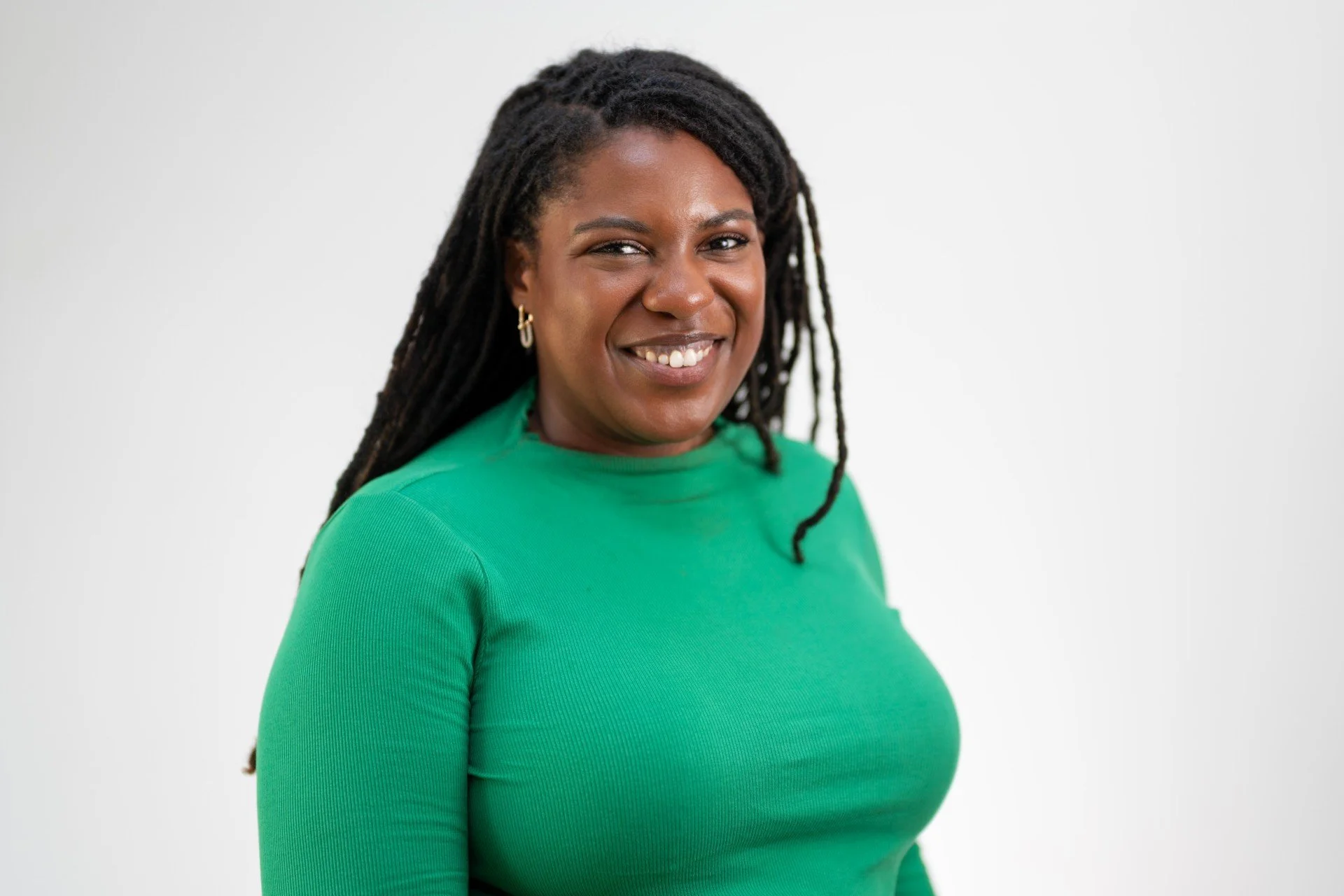 Rachel Honeyghan-Williams smiles at the camera. She's a black woman with dark skin and long dark brown locs. The background is plain and she's wearing a green t-shirt and gold earrings.
