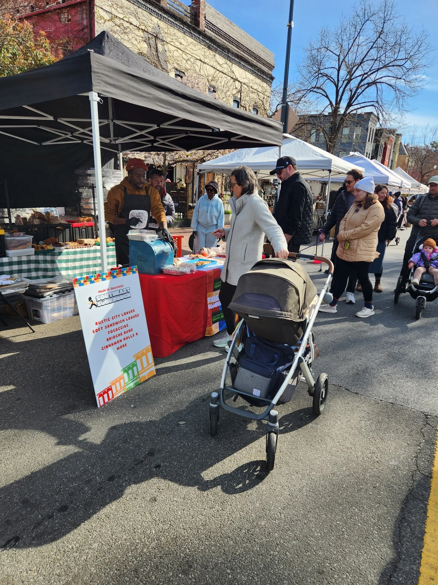 People at an outdoor market stall with a sign advertising various baked goods, including loaves of bread and cinnamon rolls, on a sunny day with trees and buildings in the background.