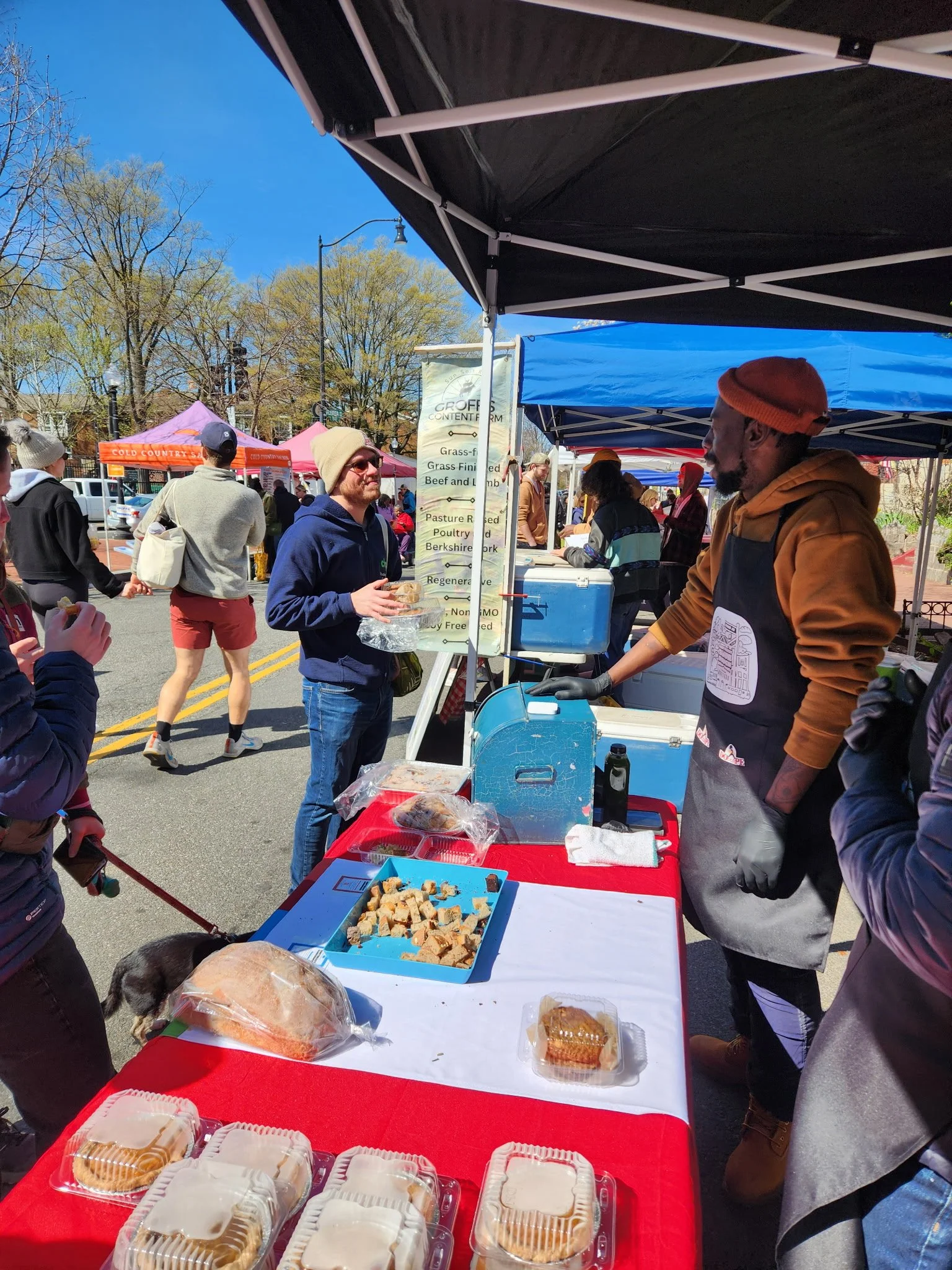 Joseph, owner of Southeast Sourdough, serving food at an outdoor market stall with baked goods, while other people browse and buy at nearby stalls on a sunny day.