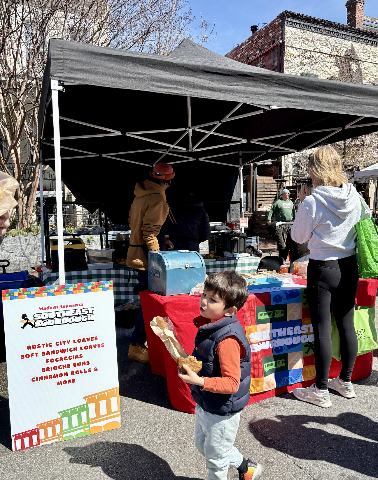 A young boy holding a piece of fried food at an outdoor market stall with a sign for Southeast Sourdough bakery. The stall sells rustic city loaves, soft sandwich loaves, focaccia, brioche buns, cinnamon rolls, and more. Several people are shopping a