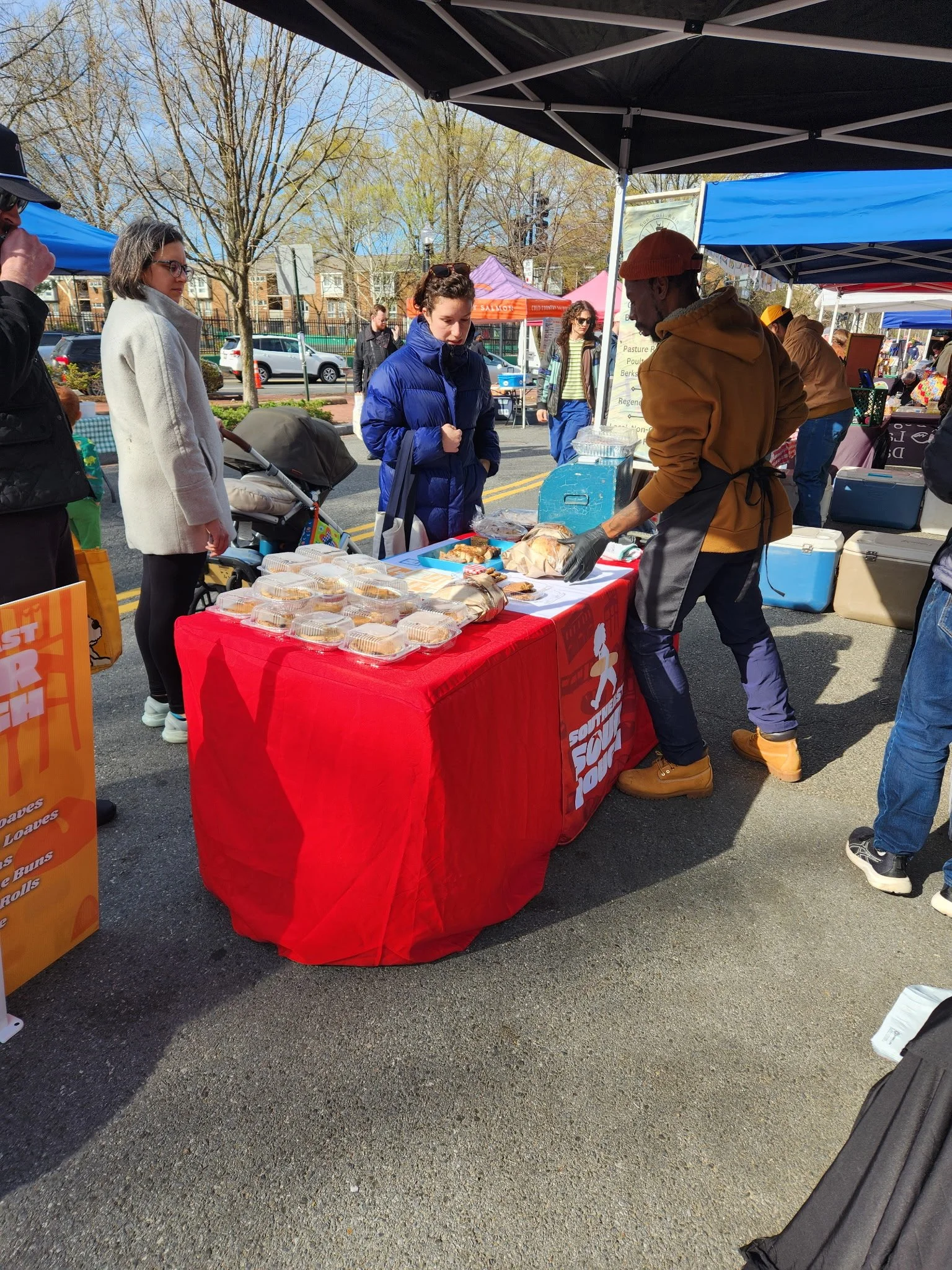 People at an outdoor farmers market with a vendor selling assorted baked goods on a Southeast Sourdough's red tablecloth.