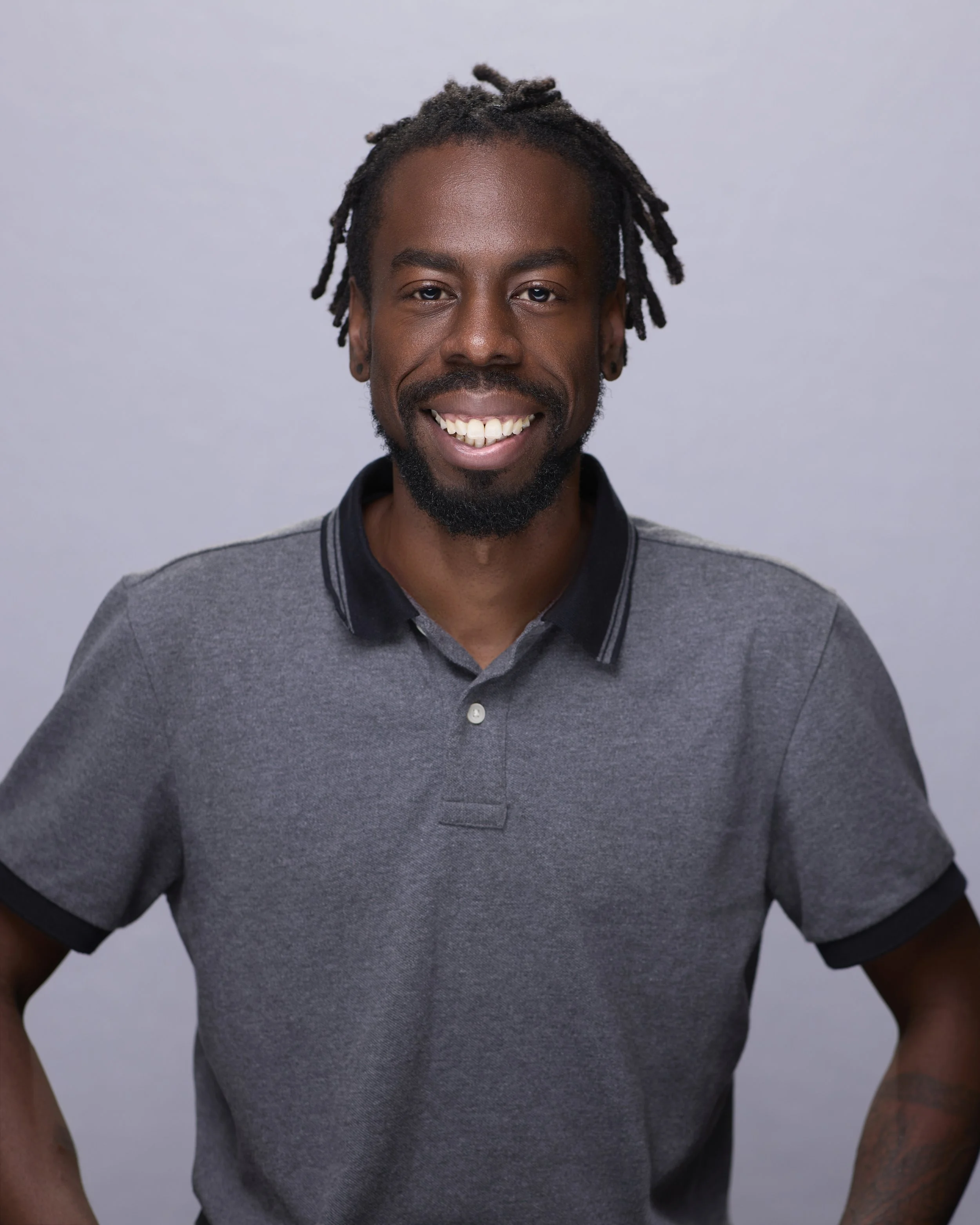 A smiling man with dreadlocks and a beard, wearing a gray polo shirt, stands against a plain light gray background.