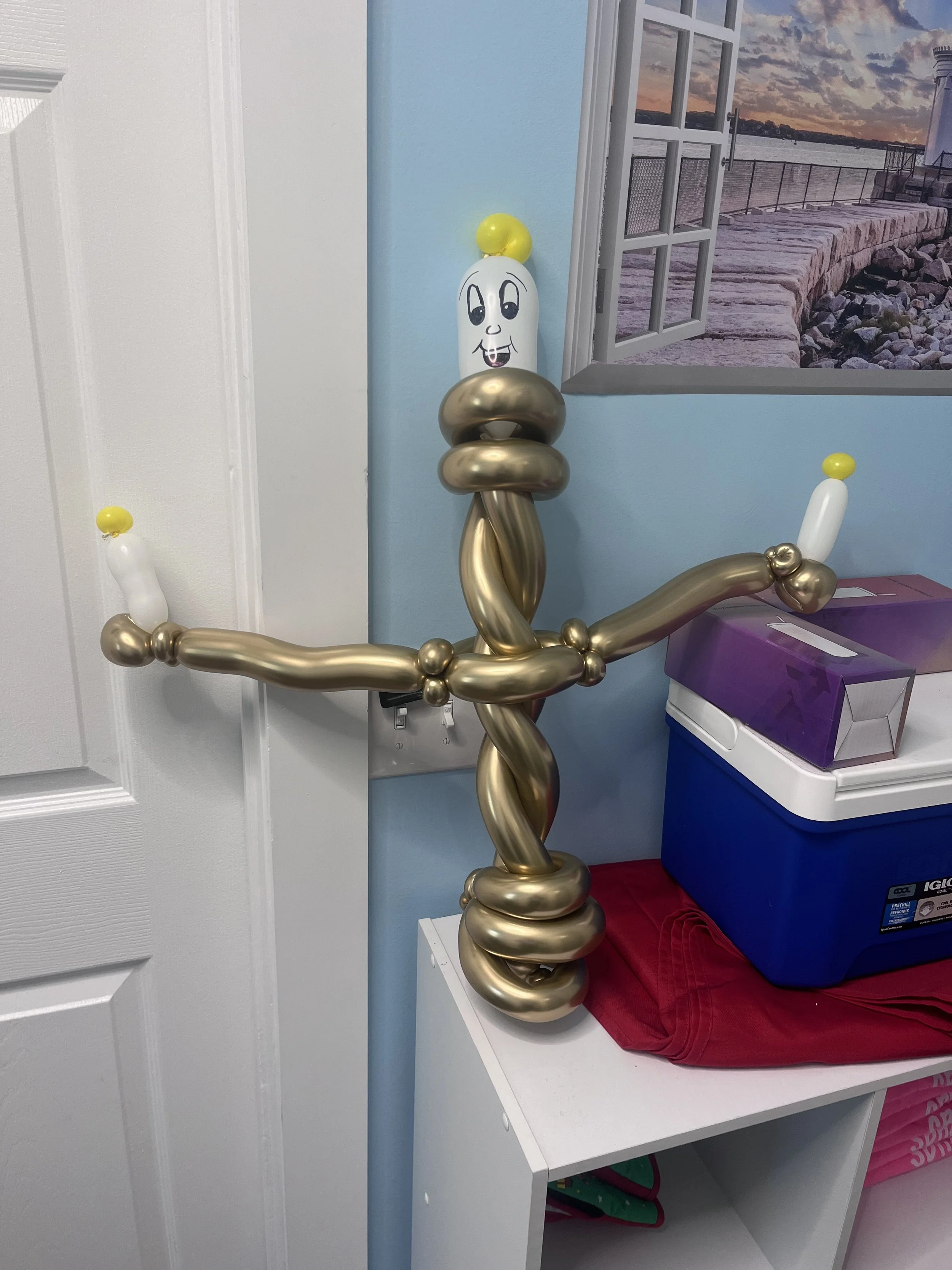 Balloon sculpture resembling a cartoonish human figure with a smiling face, arms extended outward, and a tall, twisted body, standing in a room near a door and a window.