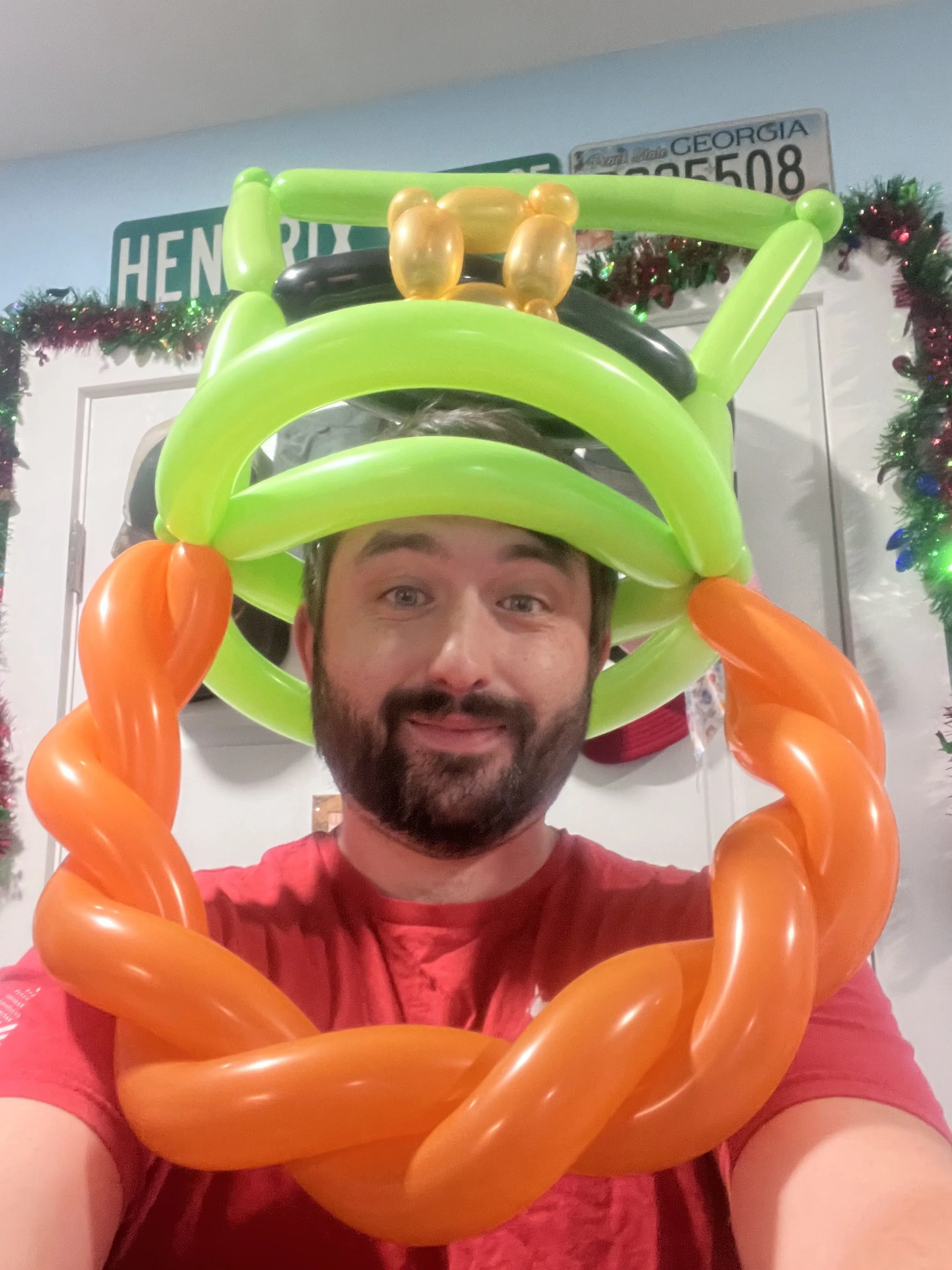A man wearing a red shirt is smiling and taking a selfie, with a colorful balloon hat featuring orange, green, black, and gold balloons, surrounded by holiday decorations on a wall.