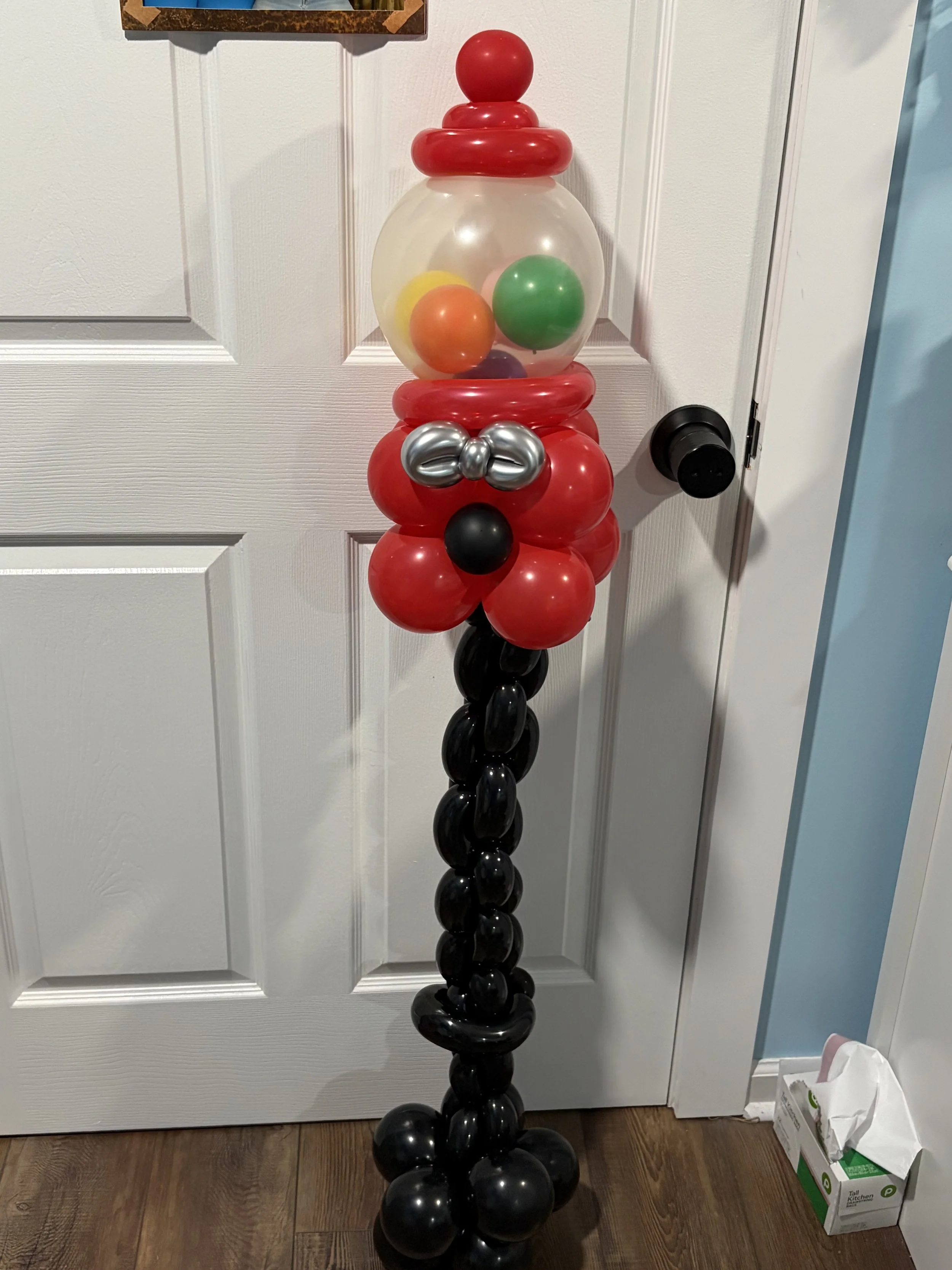 Balloon sculpture resembling Santa Claus with a transparent head filled with multicolored smaller balloons, red hat, silver bow tie, black nose, and a red suit with black balloons at the bottom as the legs, standing near a white door.