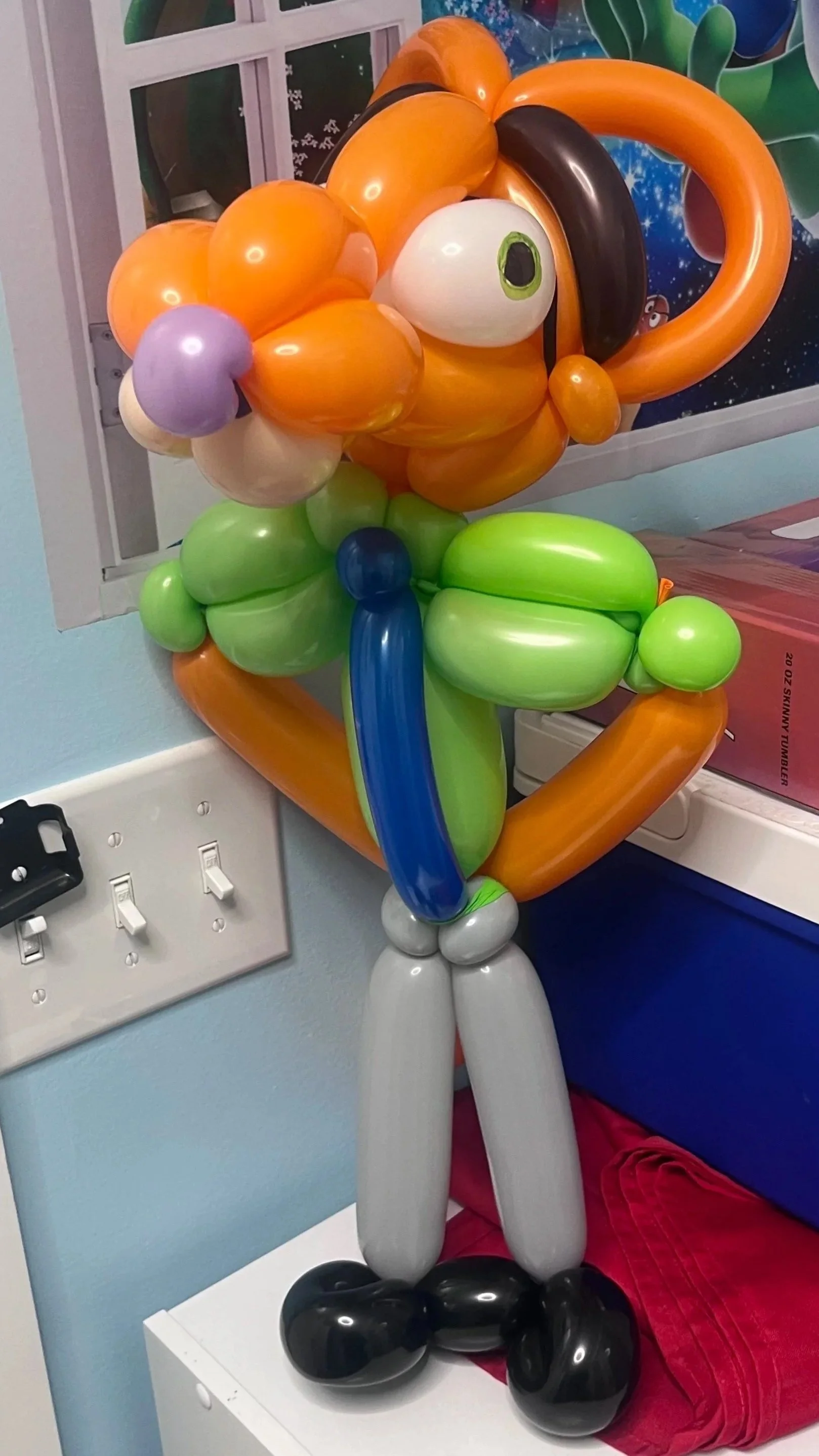 Balloon sculpture of a clown with orange hair, large eyes, green and blue clothing, and black shoes, placed on a white surface near a wall outlet.