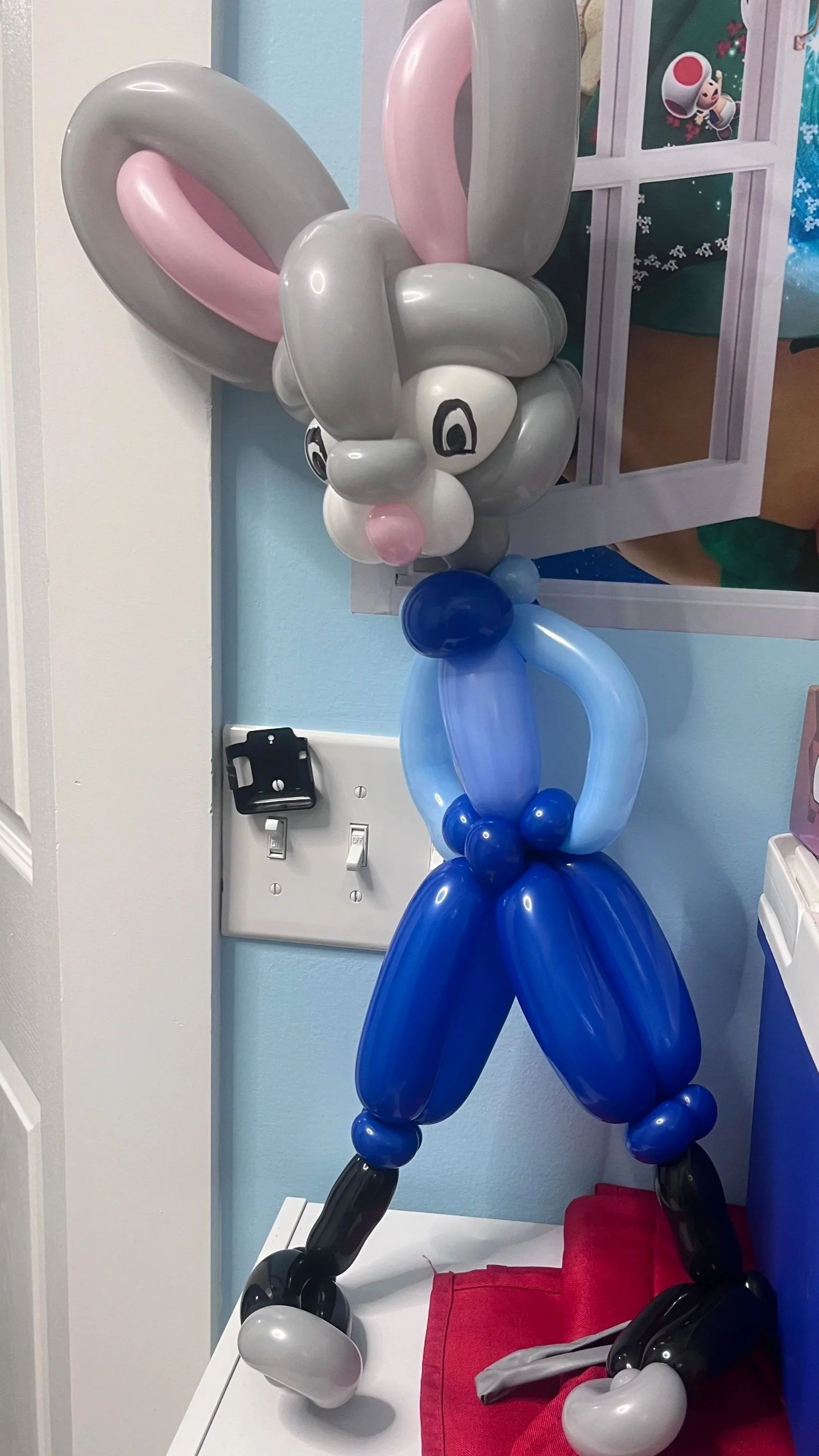 Balloon sculpture of a cartoon bunny character with pink, white, gray, blue, and black balloons, standing upright near a wall and a window with a 'Super Mario' poster.