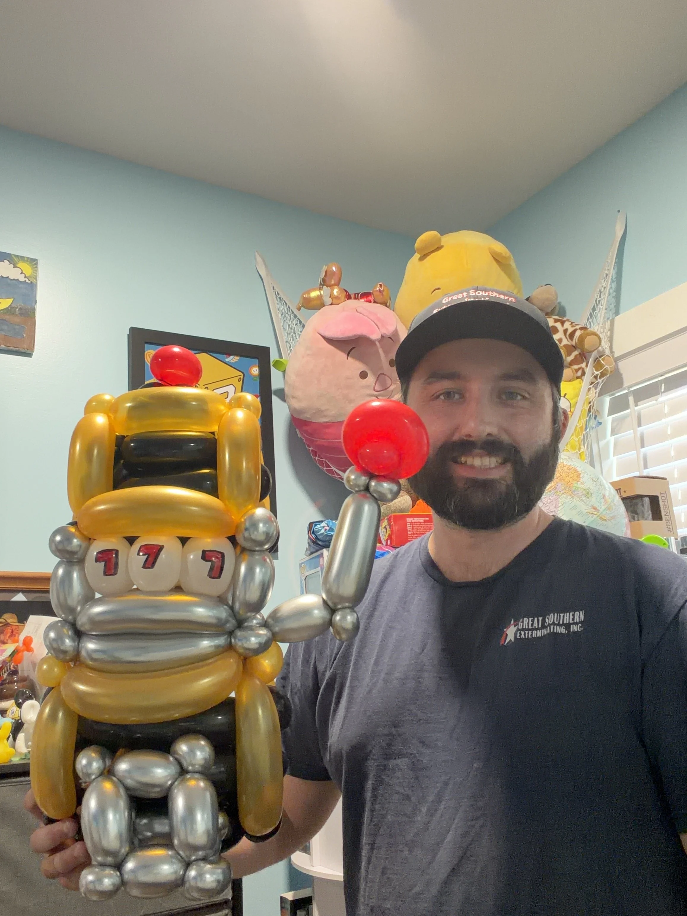 Man holding a balloon sculpture of a bee with the number 177 in a room with plush toys on the wall.