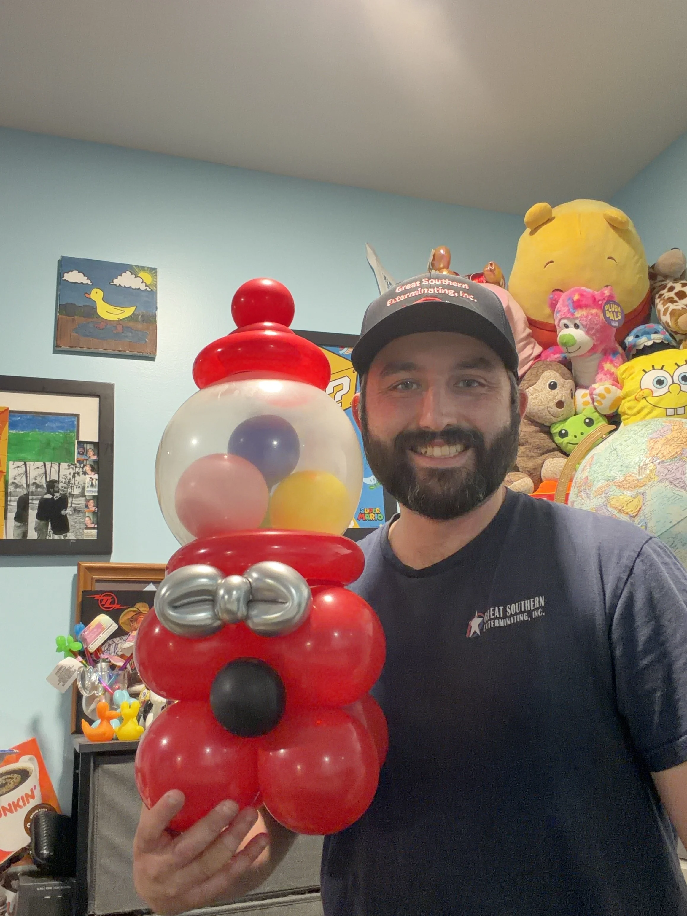 A man smiling and holding a balloon sculpture of Santa Claus in a room decorated with plush toys, framed photos, and colorful artwork.