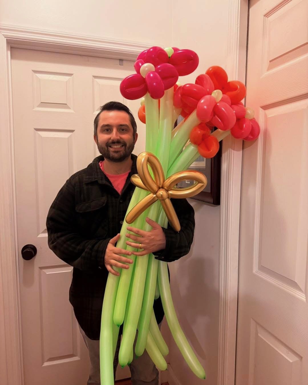 A man holding large balloon bouquet resembling flowers, made of red, orange, white, and green balloons, with a gold balloon bow in front of a white door.
