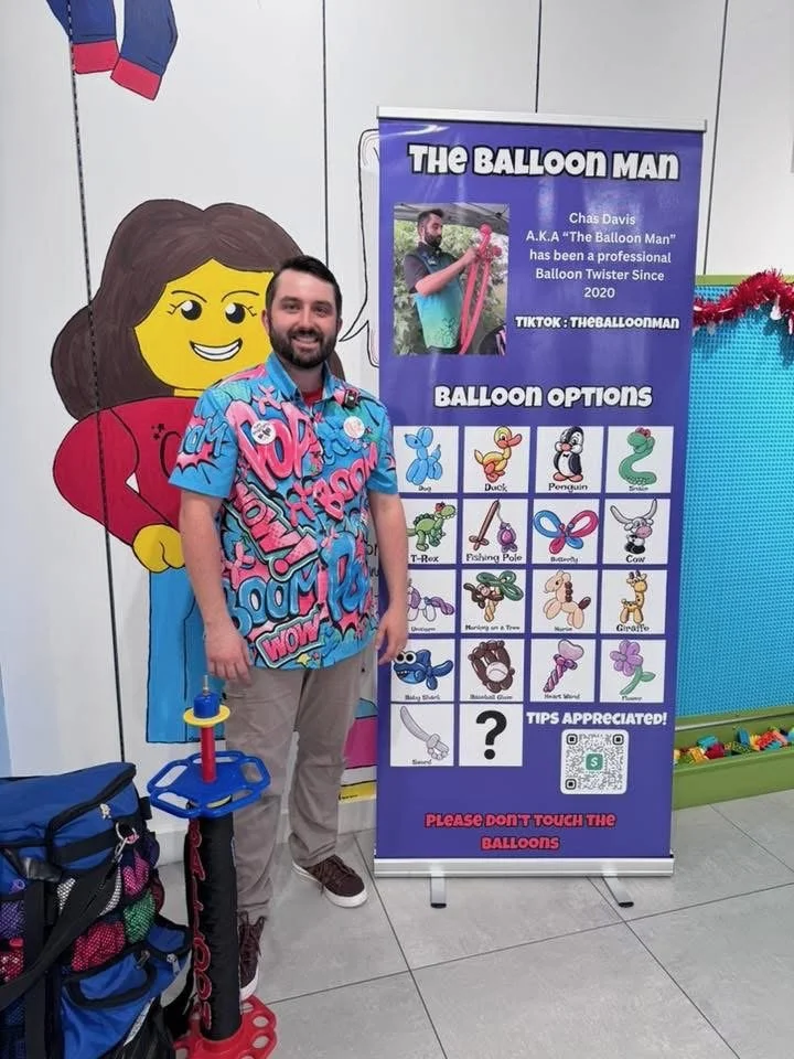 Man standing beside a balloon options chart at a fun indoor event, with a large cartoon cutout of a girl on the wall behind him and a balloon twisting tool on the floor.