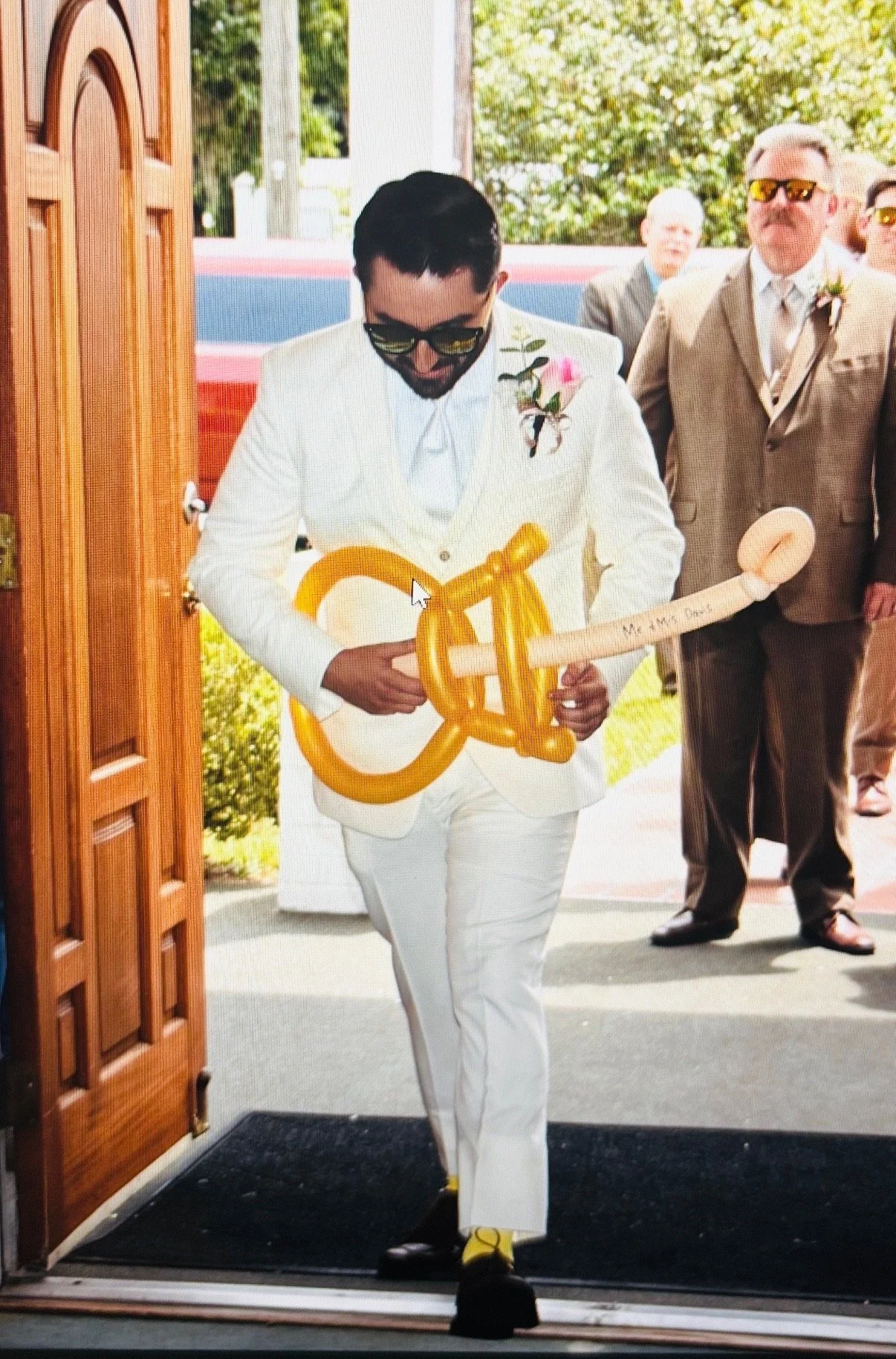 A man in a white suit holding a yellow balloon guitar with 'Mr & Mrs Dad' written on it, standing at a doorway with seated and standing people in the background. The man is wearing sunglasses and has a pink boutonniere.