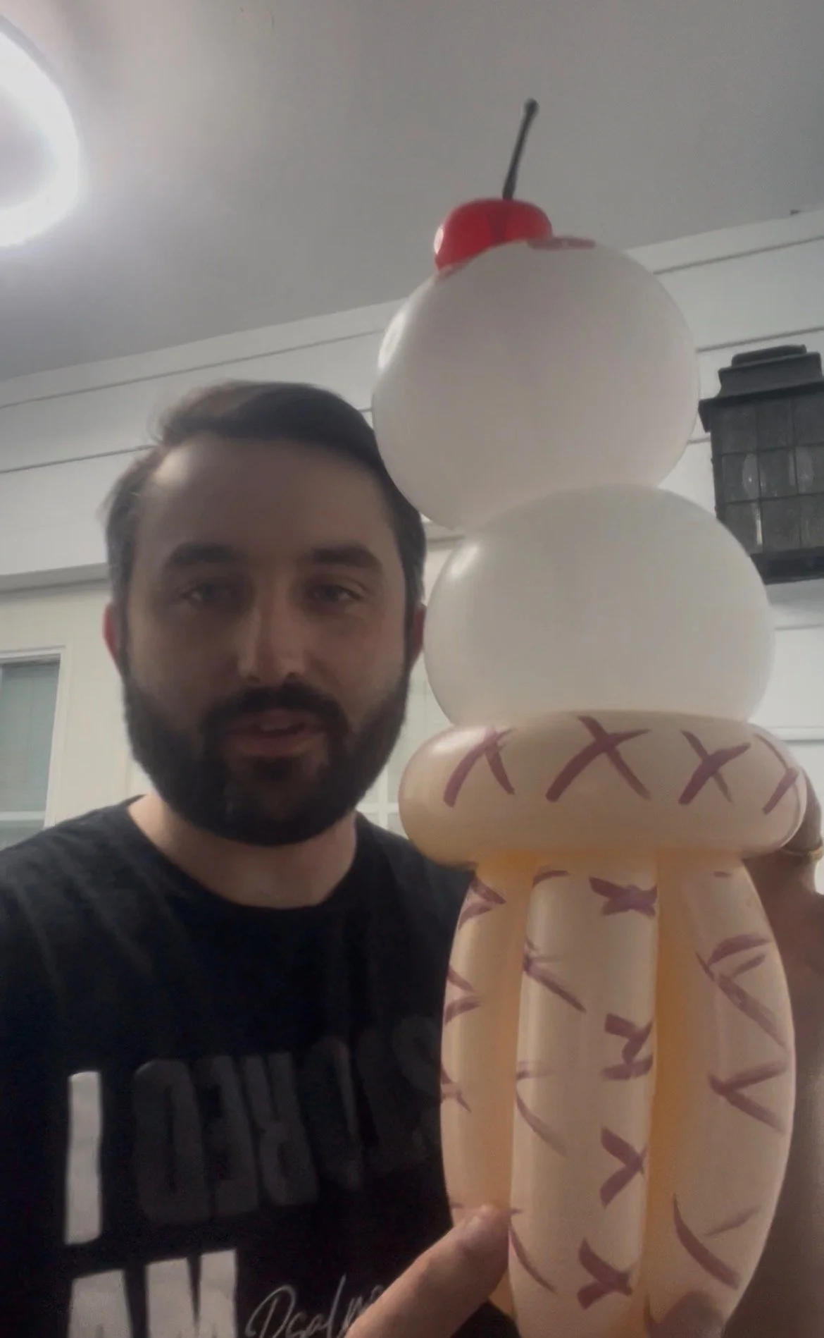 A man with dark hair and a beard holding a balloon sculpture resembling a cupcake with a cherry on top.