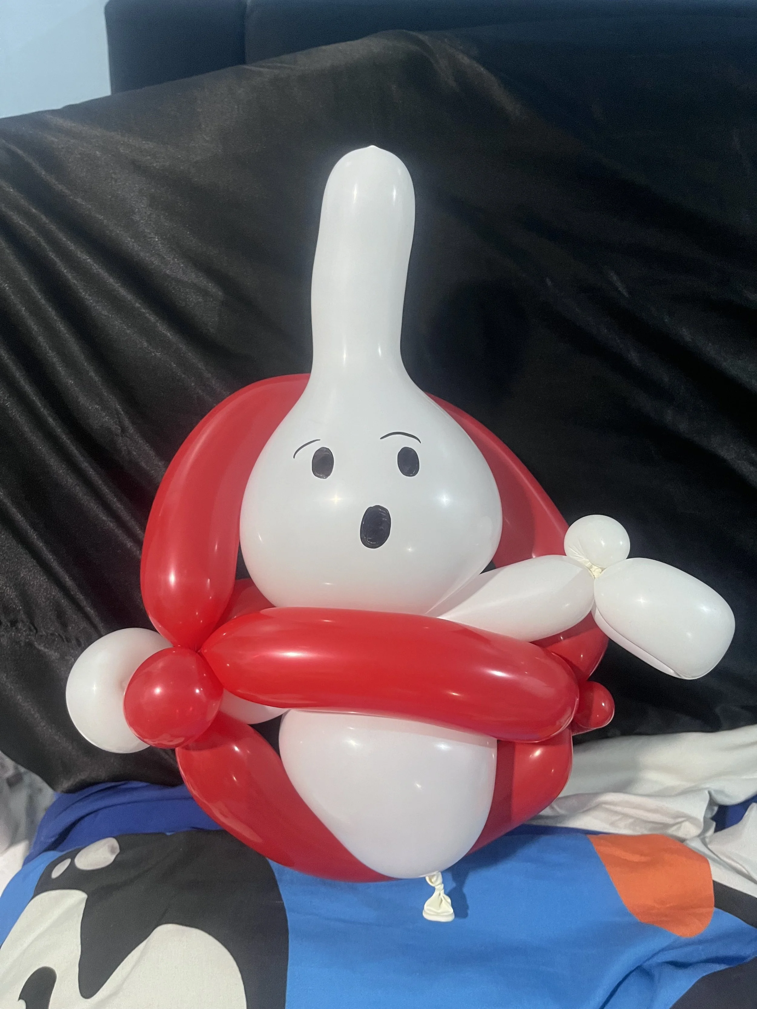 Balloon sculpture of a white cartoon ghost with black eyes and mouth, surrounded by red balloons shaped like a ghost's cape or cloak, placed on a surface with a black fabric background.