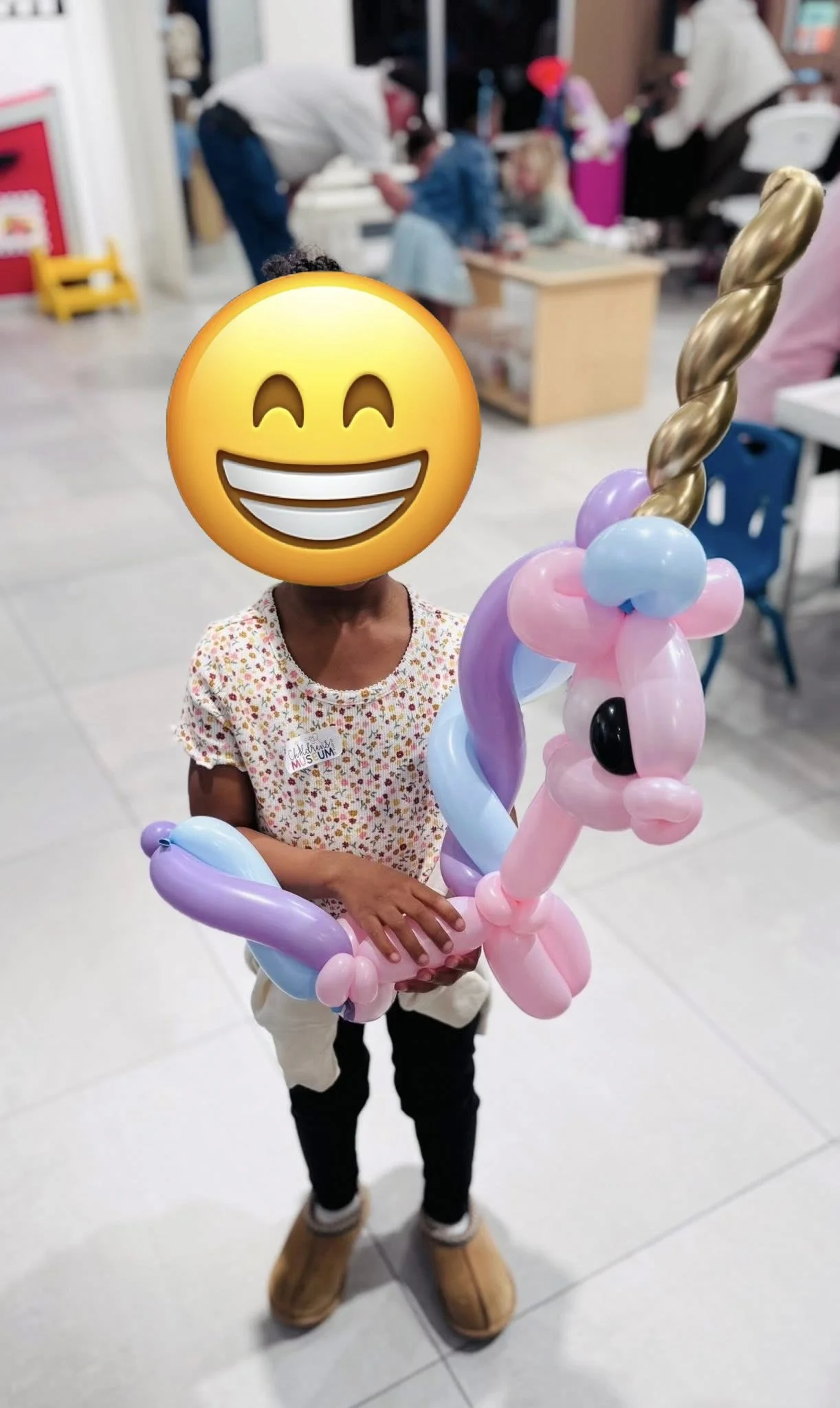 A child holding a pink, purple, and blue balloon unicorn with a golden twisted balloon horn, in what appears to be a toy or shopping store.