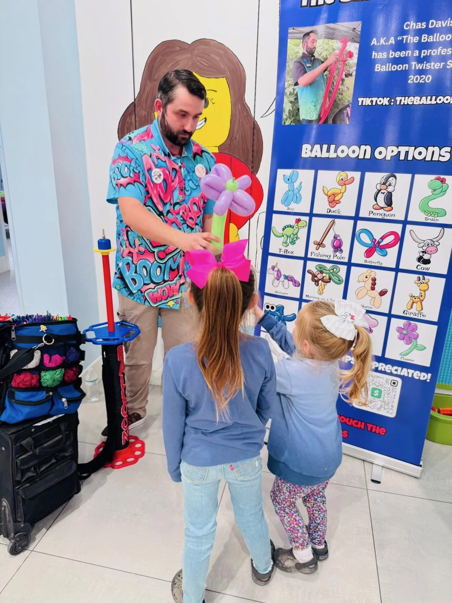 A man in a colorful shirt makes balloon animals for two young girls at a balloon art booth. The booth features a blue poster with balloon options and a picture of the balloon artist.