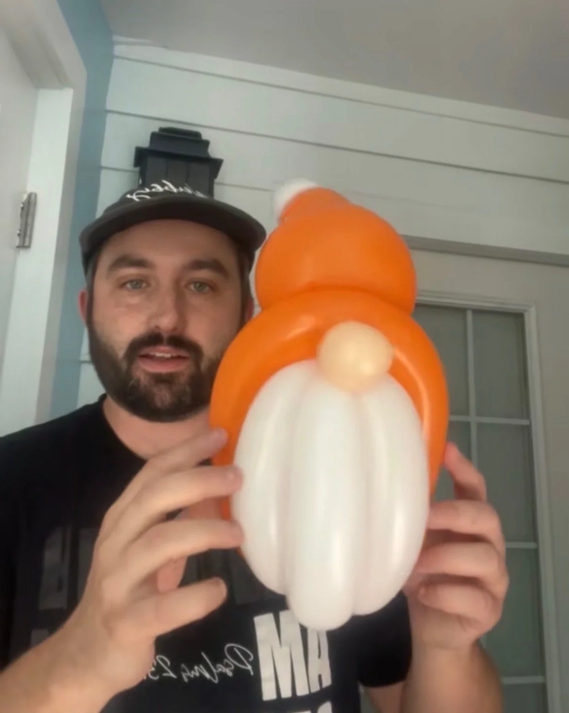A man with a beard, wearing a black shirt and a gray cap, holding a balloon sculpture resembling a snowman, made of orange, white, and beige balloons, indoors with white and blue trim walls.