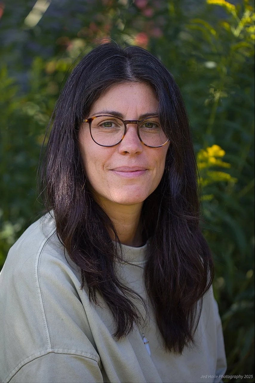 A woman with dark hair and glasses smiling outdoors with greenery in the background.