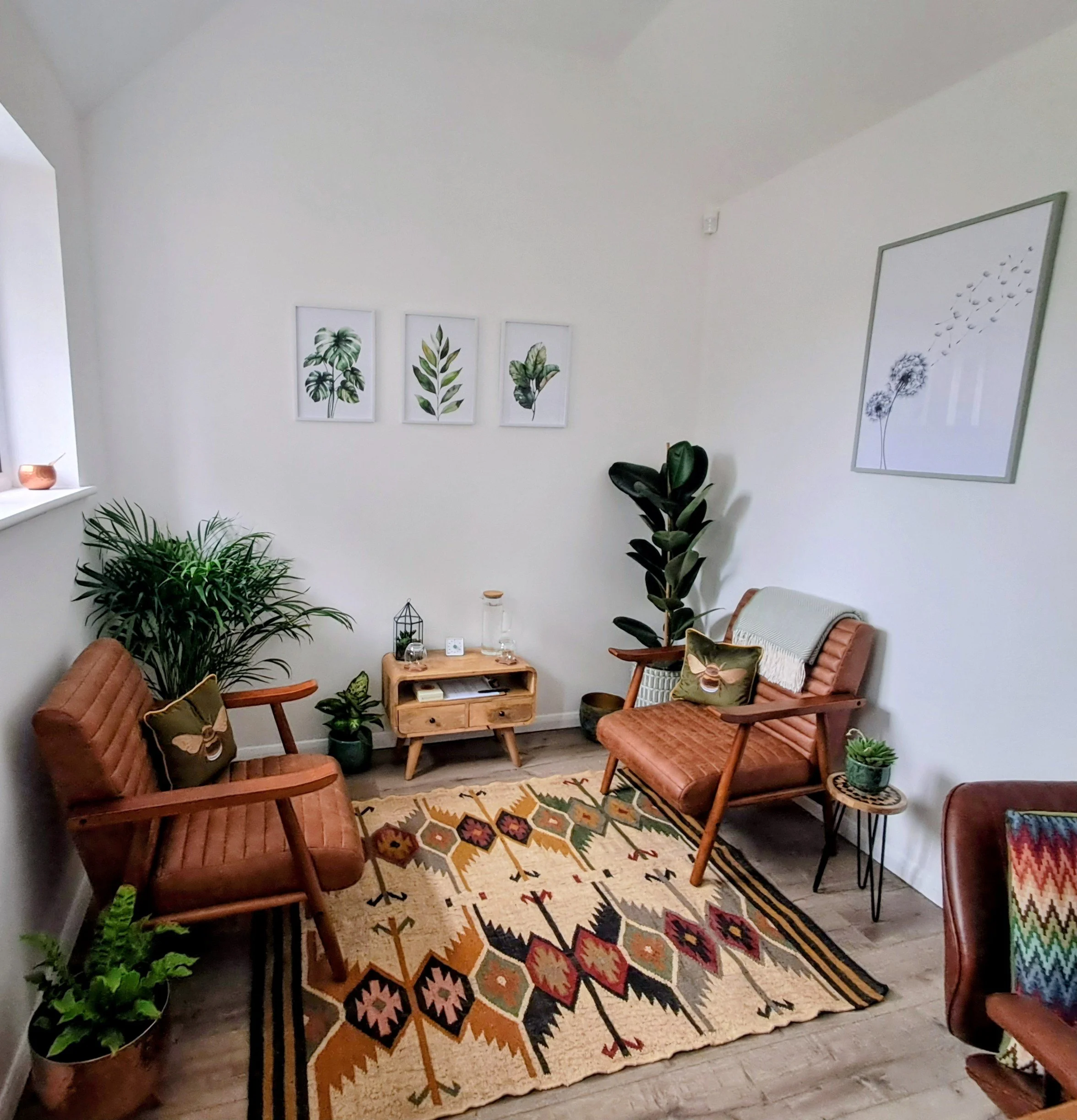 Living room decorated with plants, framed botanical art, and colorful geometric-patterned rug. Wooden furniture includes two chairs and a small table with decorative items.