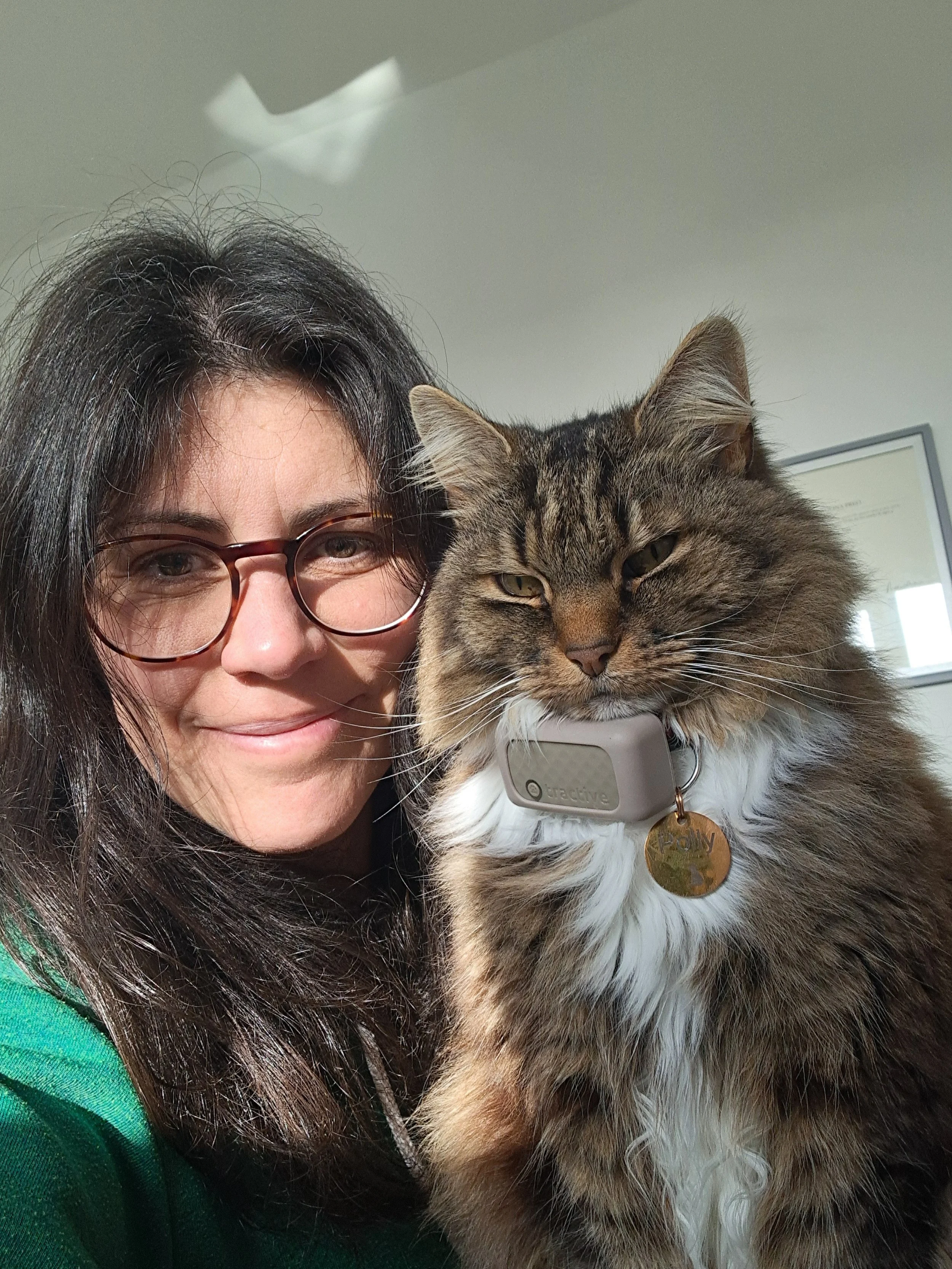 A woman with long dark hair and glasses smiling next to a large tabby cat with a collar and a gold tag, indoors with a framed picture on the wall in the background.