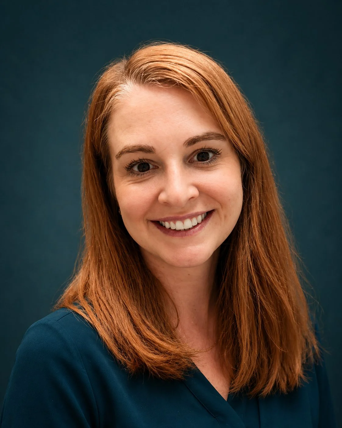 A portrait of a smiling woman with red hair, wearing a navy blue top, against a dark teal background.