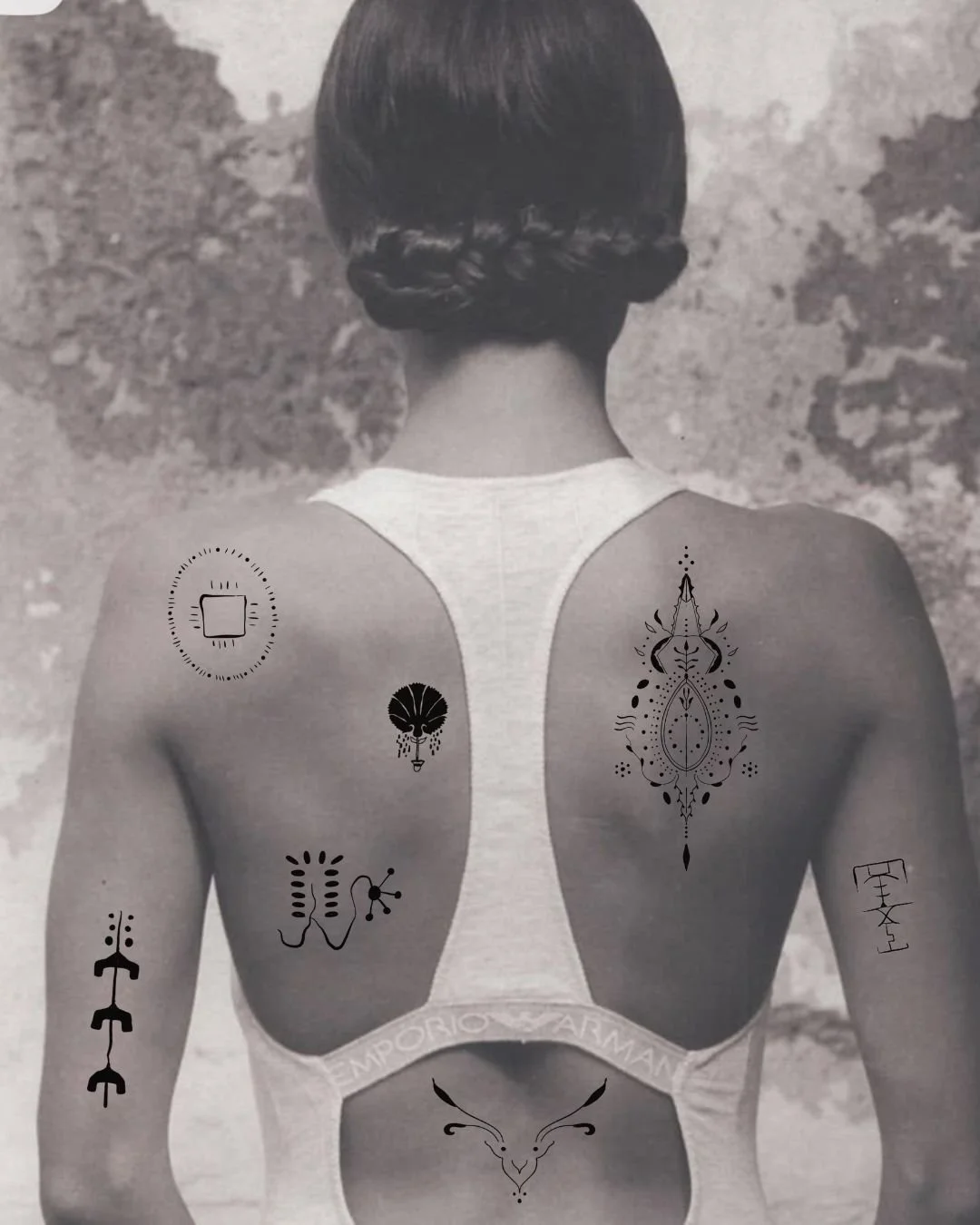 A woman with her back to the camera, showcasing various black tattoo designs on her arms and upper back, wearing a white sports bra, with a textured wall in the background.