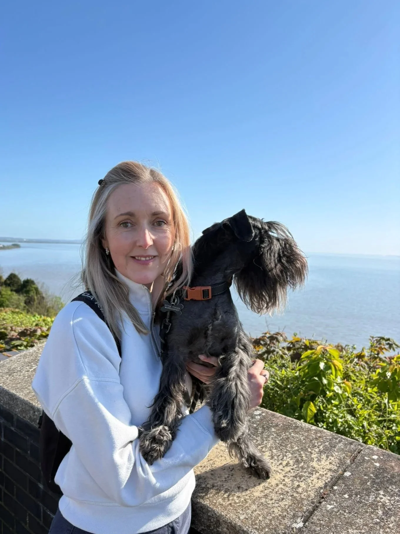 A woman with blonde hair, wearing a white jumper, holding a black and miniature schnauzer dog with long ears and a collar, standing outdoors overlooking the sea, with clear blue sky and greenery in the background.