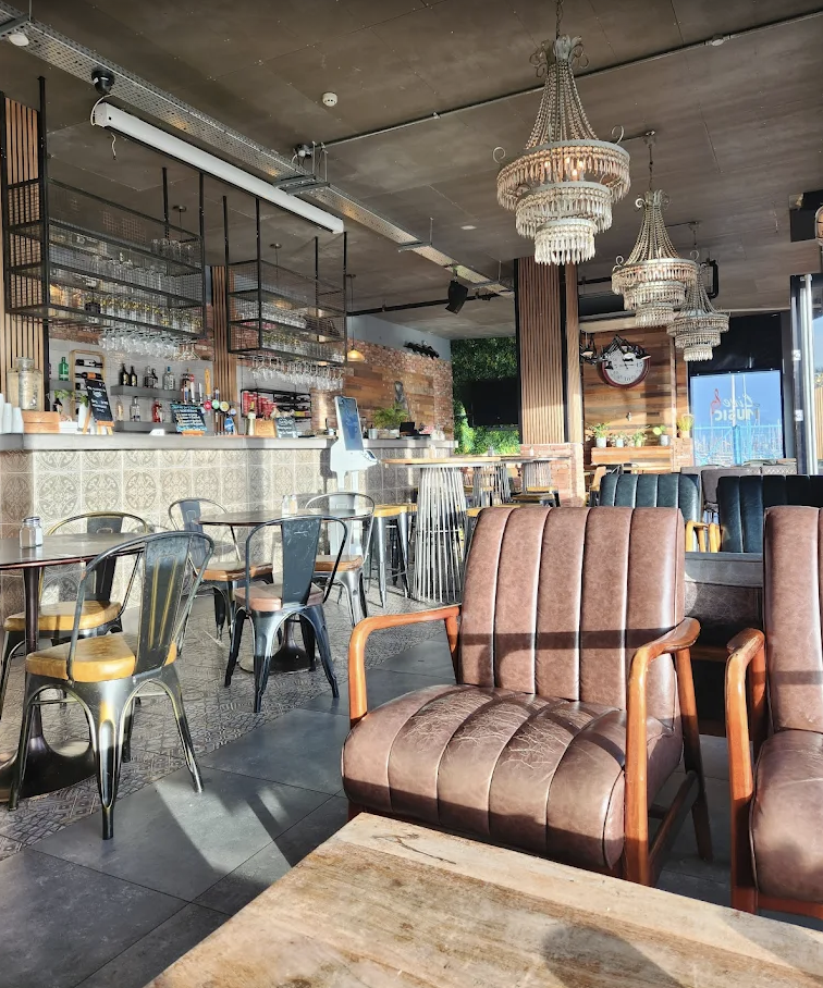 Interior of a Coffi Co at Bayscape, Penarth, with vintage-style chairs, chandeliers, a bar area with glasses, bottles, and a digital ordering kiosk, and a large window showing outside greenery.