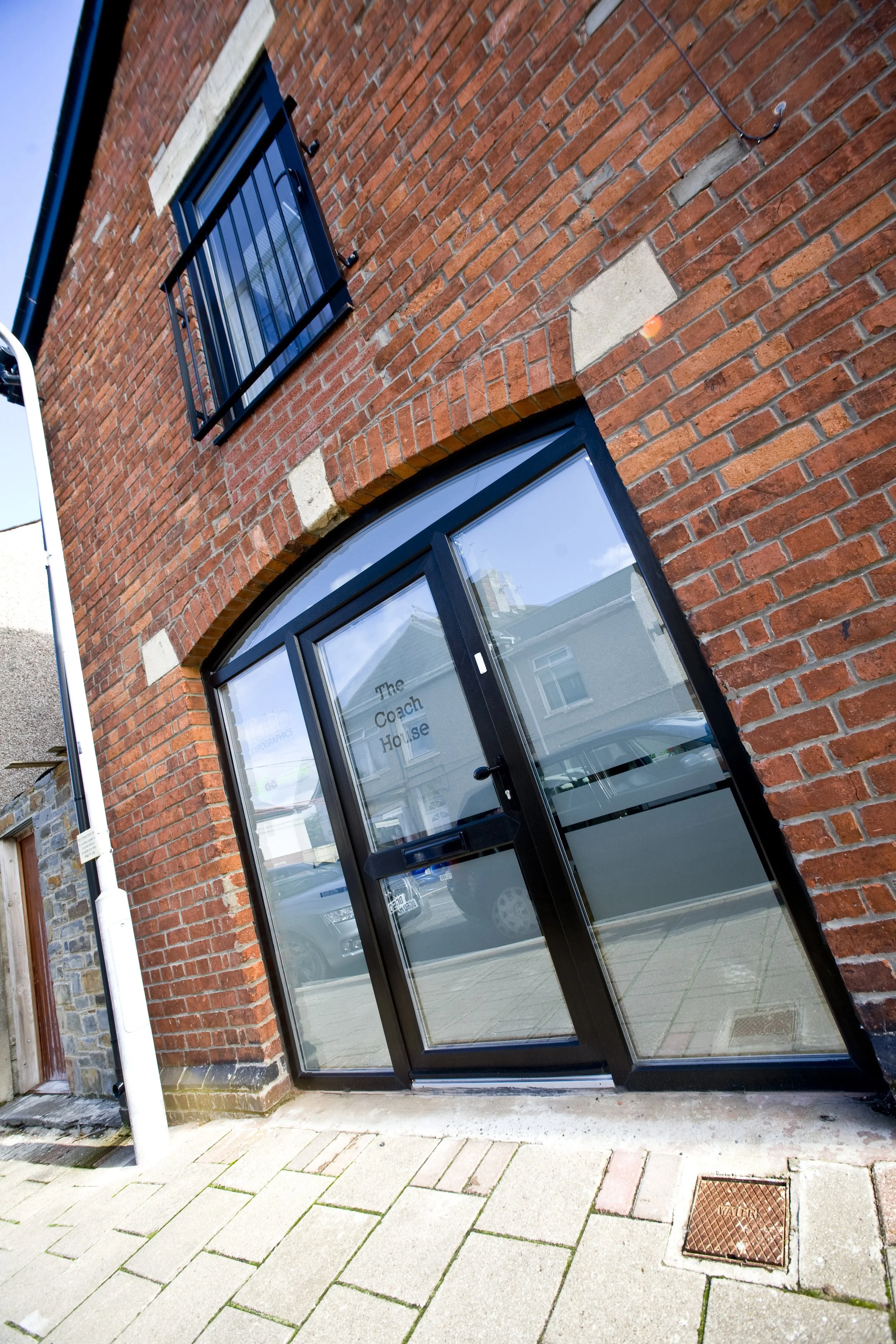 Red brick building with a black-framed glass door and a small black window with bars, labeled "The Coach House." Paved sidewalk in front, with a drain cover and a white pole on the sidewalk. Reflection of neighboring buildings and cars in the glass door.
