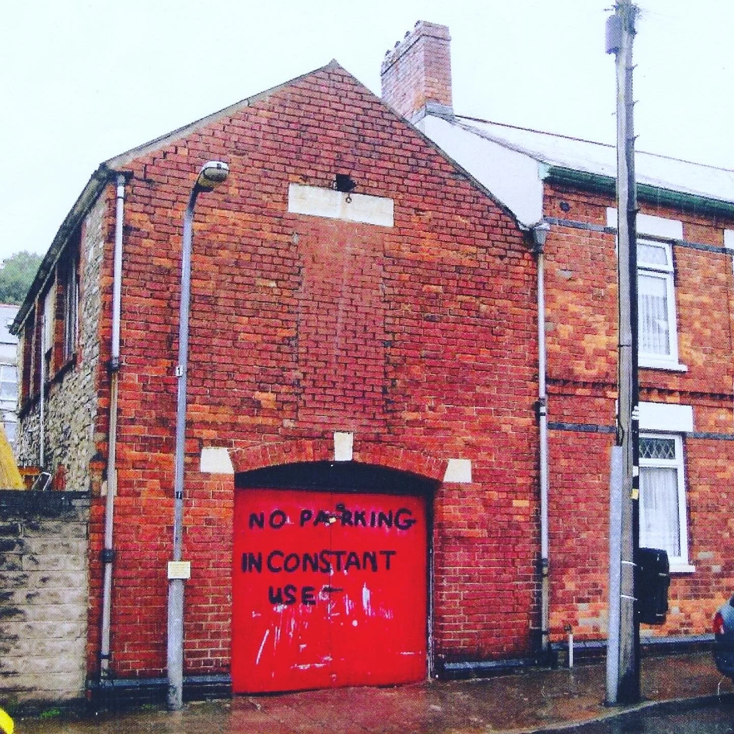 Red brick building with a painted red garage door. The building has a sloped roof, windows, and drainage pipes visible, with overcast weather.