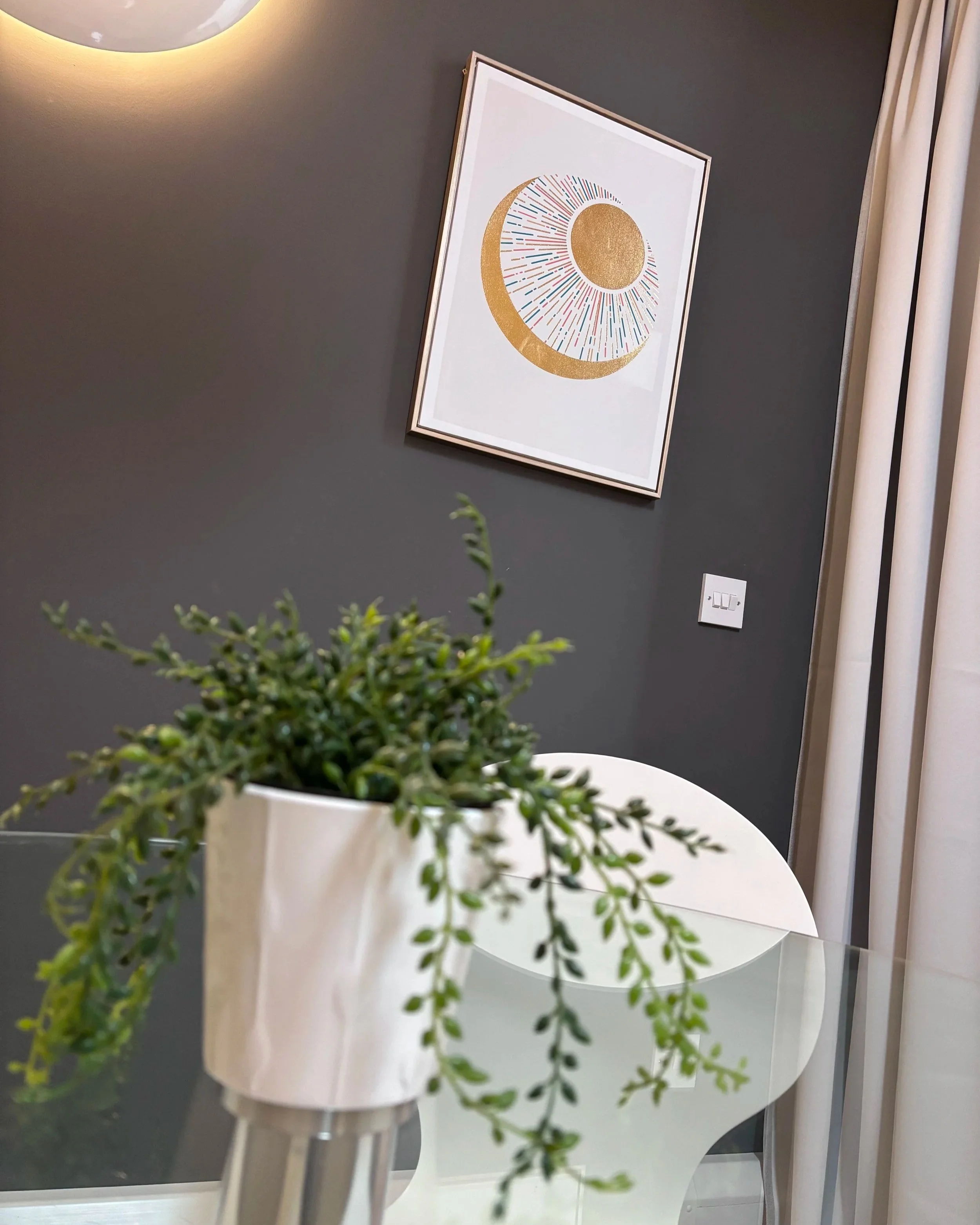 A framed art print with a gold and multicolored design hangs on a dark gray wall near cream curtains. A white table with a potted plant is in the foreground.