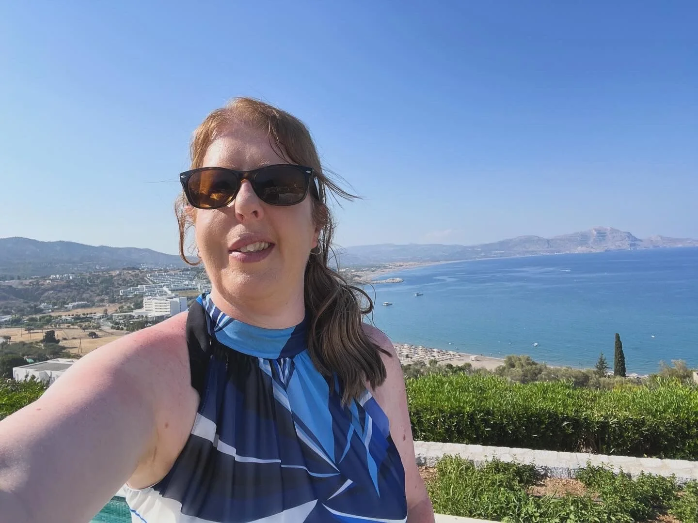 A woman taking a selfie outdoors with a coastal view of blue ocean, mountains, and clear sky in the background.