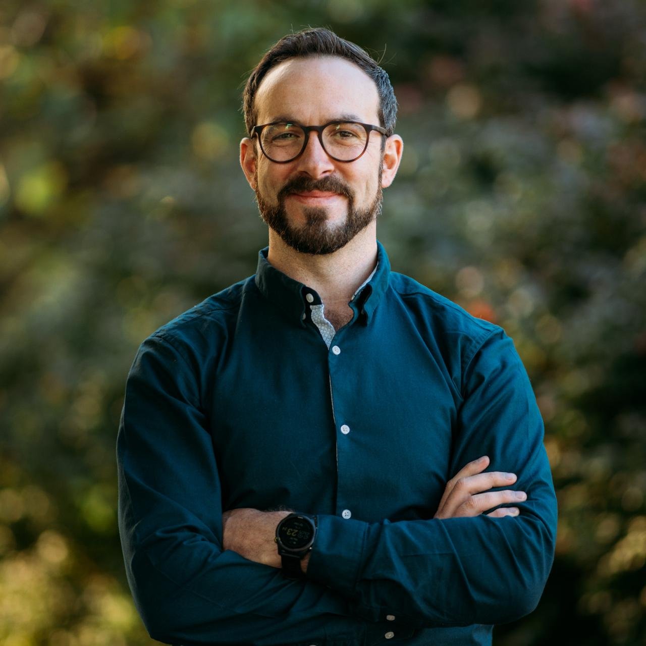 A man with glasses and a beard smiling, standing outdoors with his arms crossed, wearing a dark green button-up shirt and a smartwatch, with blurred trees in the background.