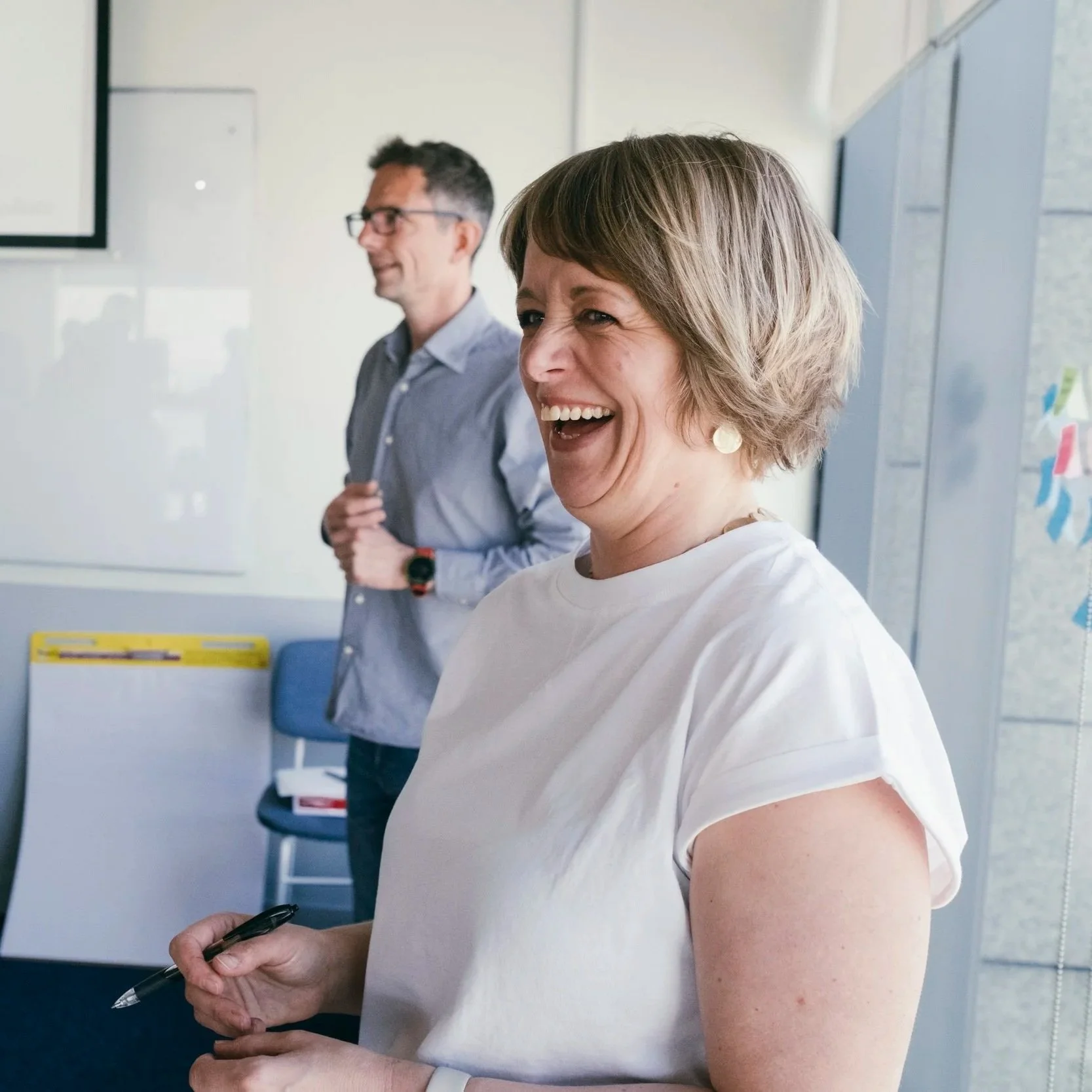 A woman with short brown hair and earrings laughs in a workroom, standing near a whiteboard and large windows. A man with glasses and dark hair, wearing a blue shirt, stands in the background.